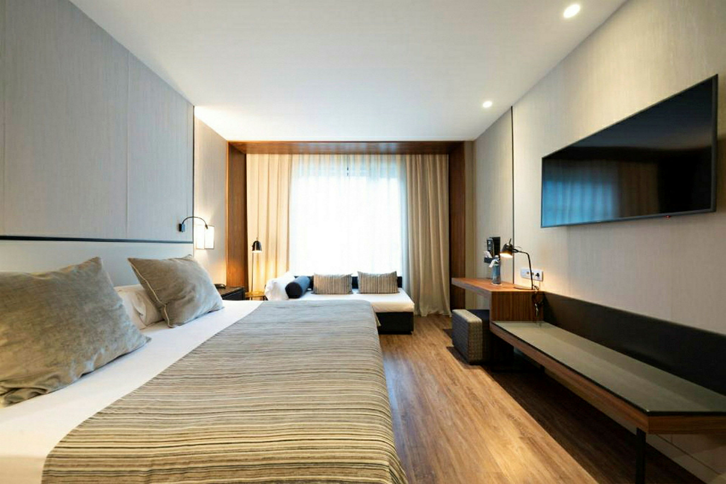 Et hotellrom med en stor seng, tilhørende nattbord og lamper samt en sittegruppe med sofaputer. Rommet har tregulv og en veggmontert TV. (AI-generert tekst)