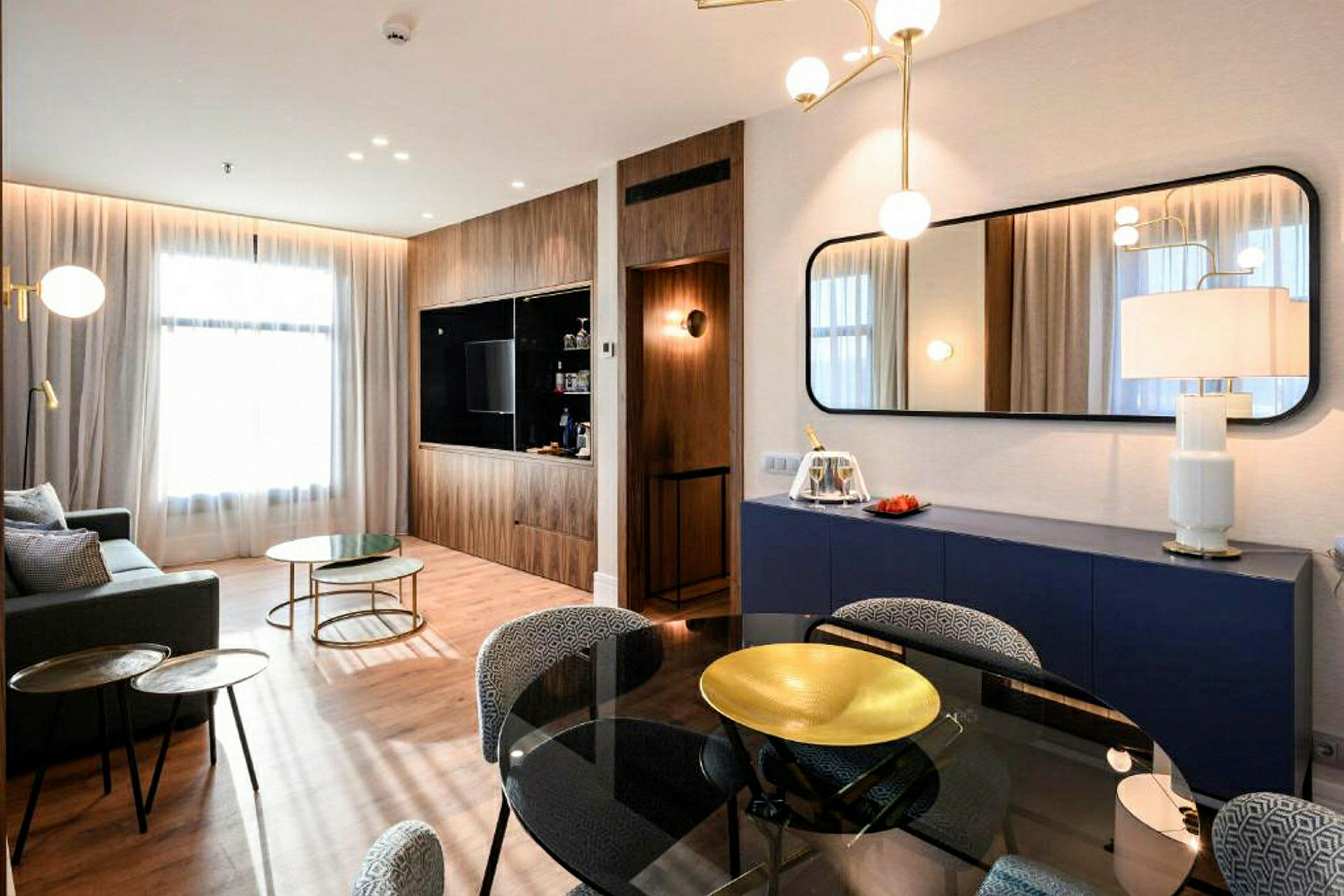 Et moderne innredet stue med sofa, glassbord og stoler. En TV er montert på en vegg med trepanel, og et stort speil henger over en blå oppbevaringsmøbel. (AI-generert tekst)