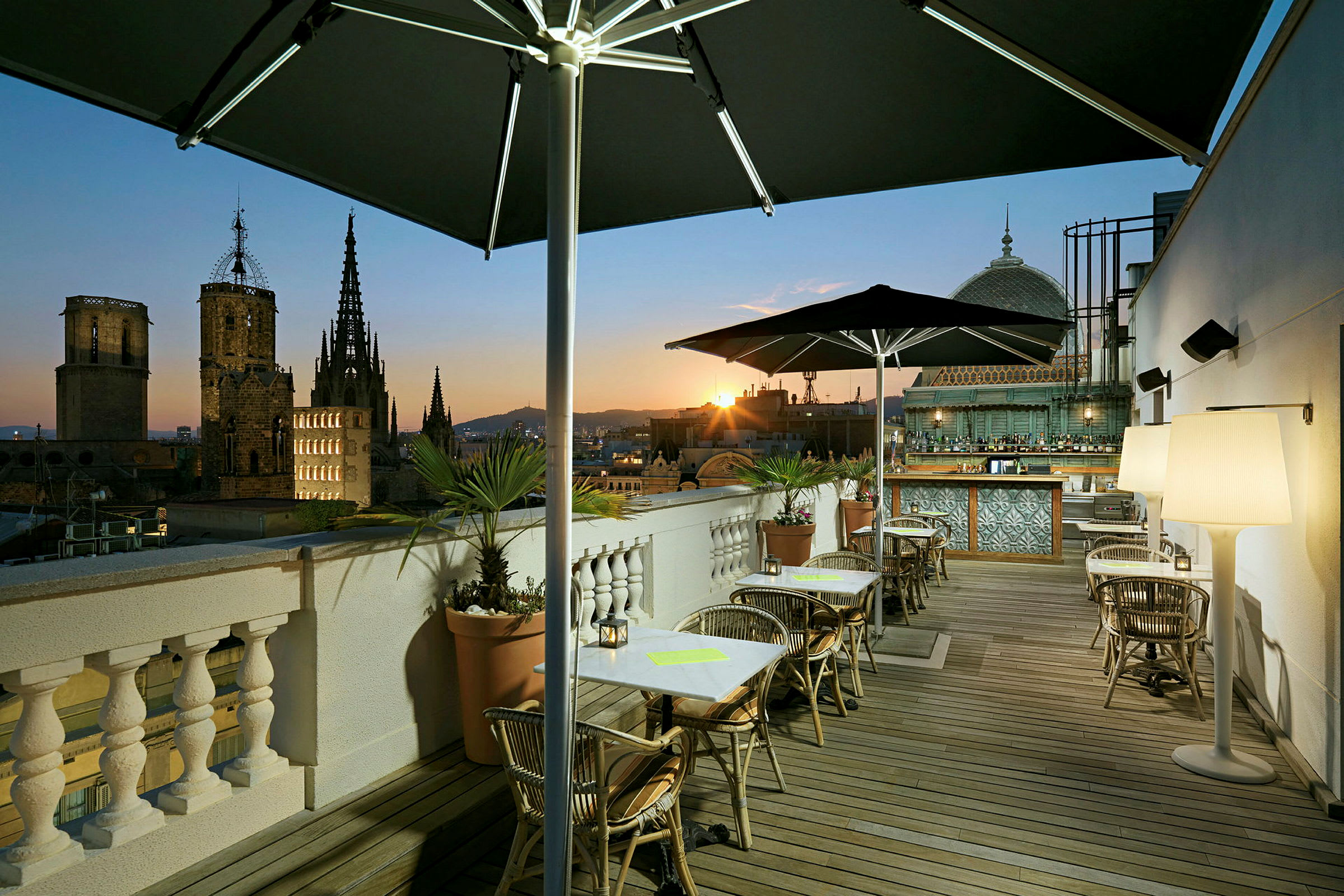 En takterrasse med bord og stoler, parasoller og planter, med utsikt over en by med katedral i solnedgangen. (AI-generert tekst)