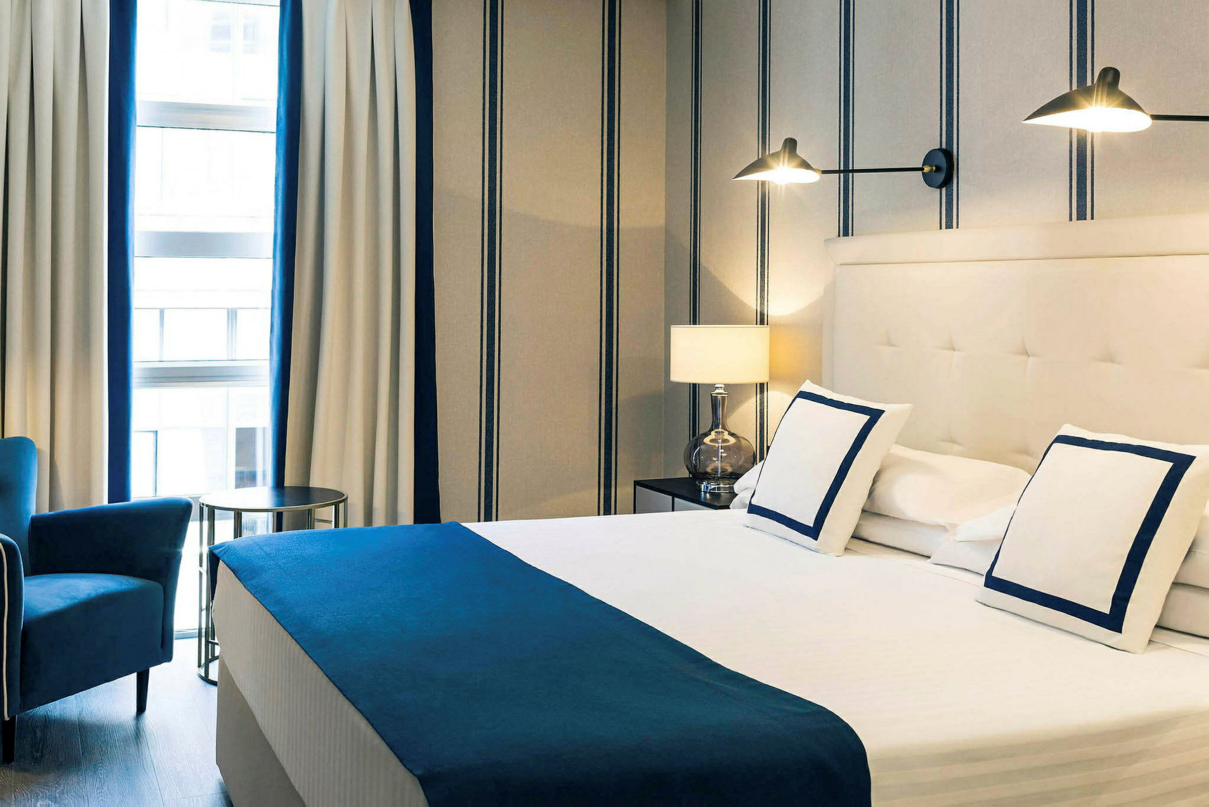 Et hotellrom med en dobbeltseng, hvitt sengetøy med blå detaljer, en lenestol og stripete vegger og gardiner. (AI-generert tekst)