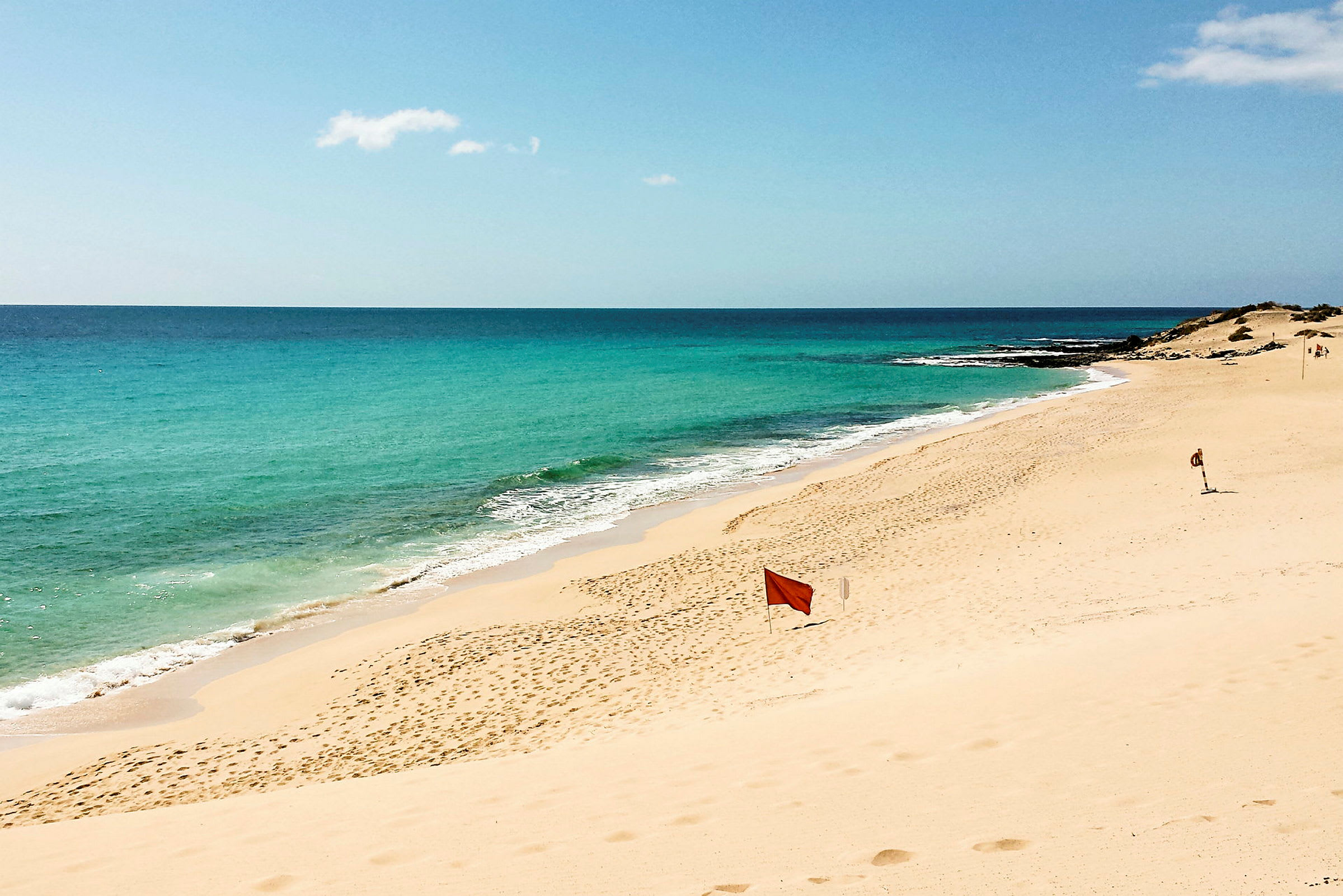 En sandstrand med turkis vann og blå himmel. Et rødt advarselsflagg er plassert på stranden. (AI-generert tekst)