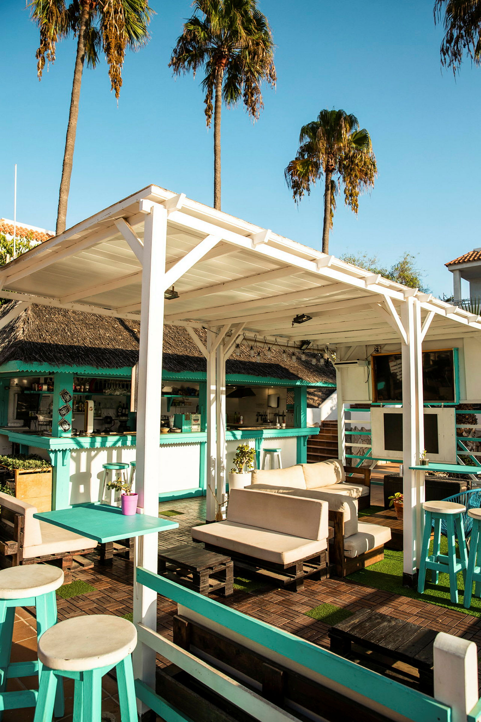 En utendørs lounge med sofaer og bord under en hvit trepergola, omgitt av palmer og en bar. (AI-generert tekst)