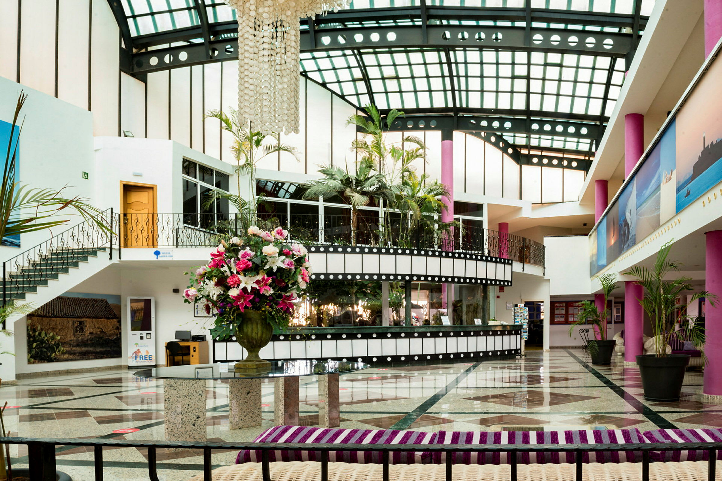 Interiør av en hotellobby med glasstak, rosa søyler og en stor vase med fargerike blomster. (AI-generert tekst)