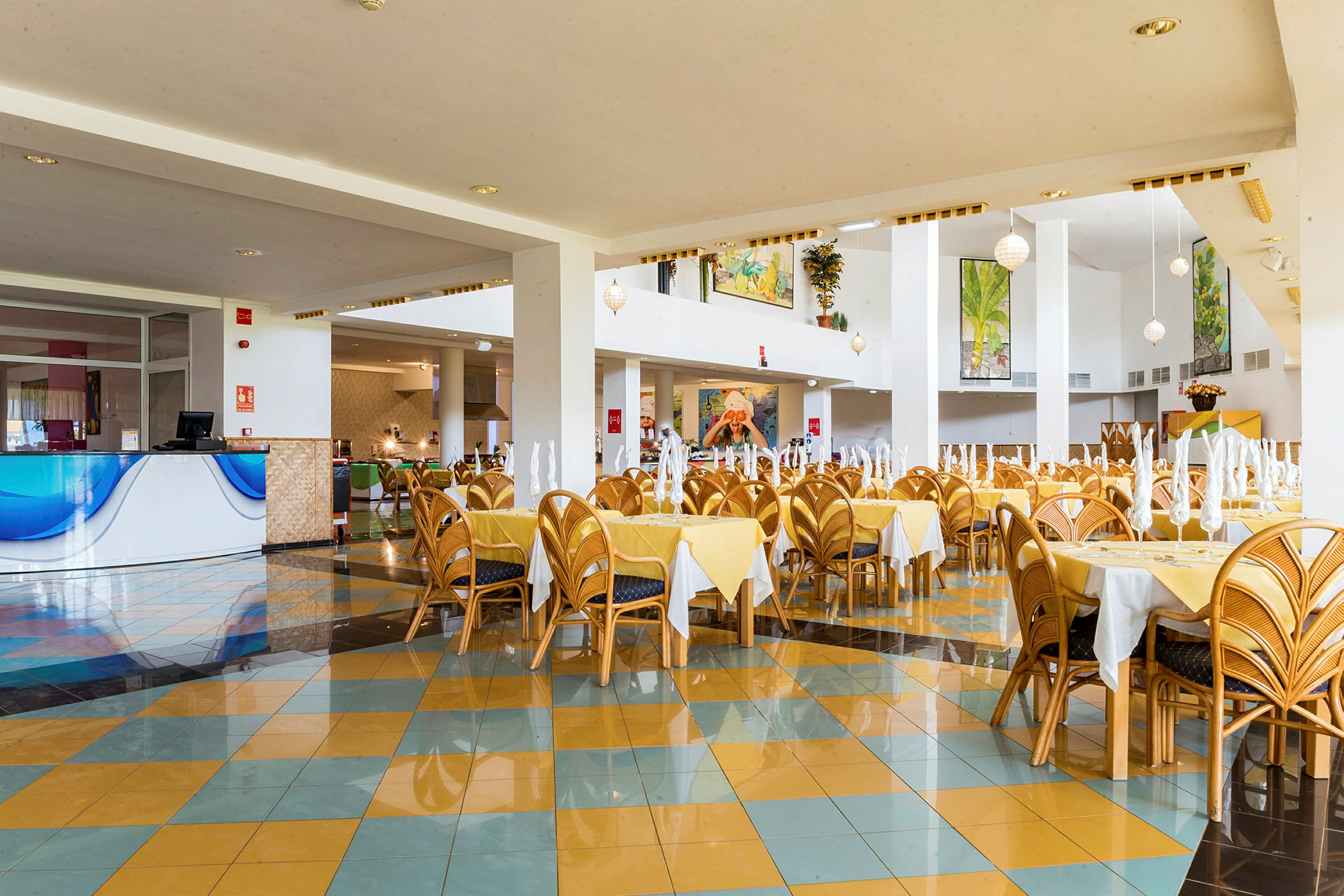 En romslig restaurant med gule duker og stoler av tre. Flere bord er dekket med brettede servietter, og lokalet har fine detaljer og dekorasjoner. (AI-generert tekst)