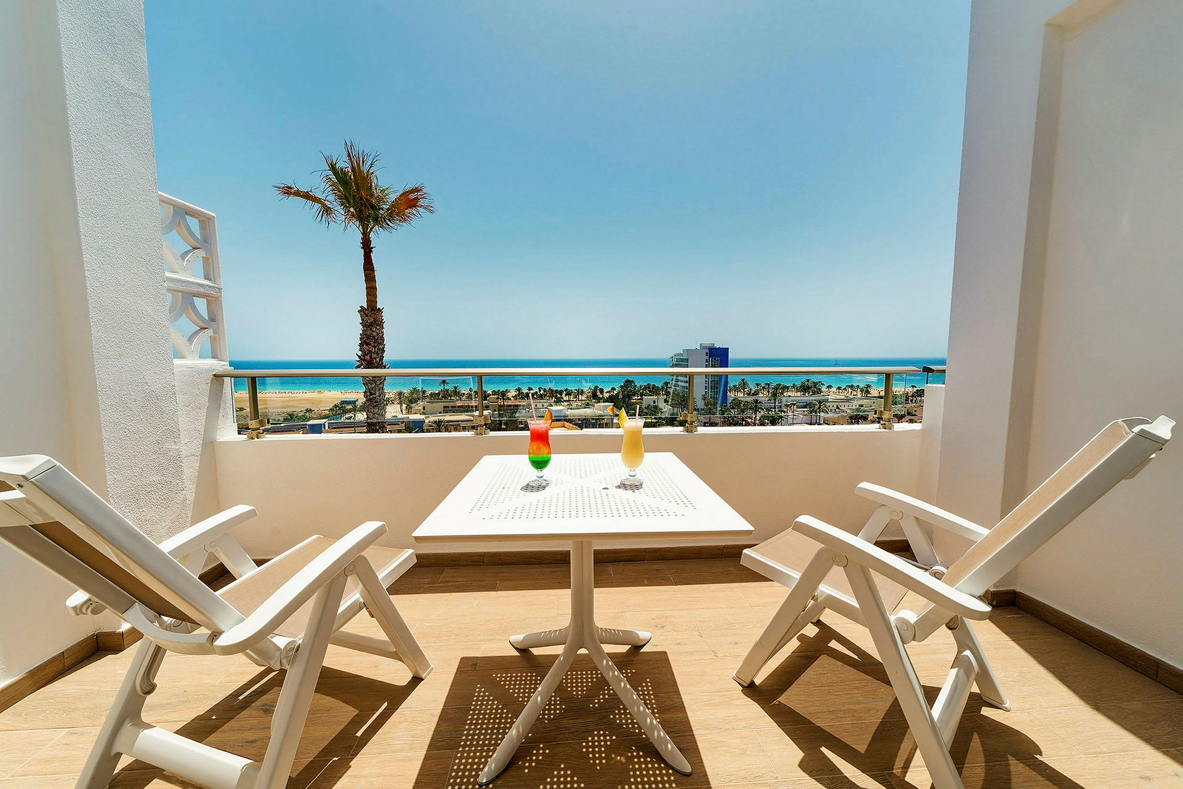 Terrasse med to hvite stoler og et bord, to drinker og utsikt over stranden og havet. (AI-generert tekst)
