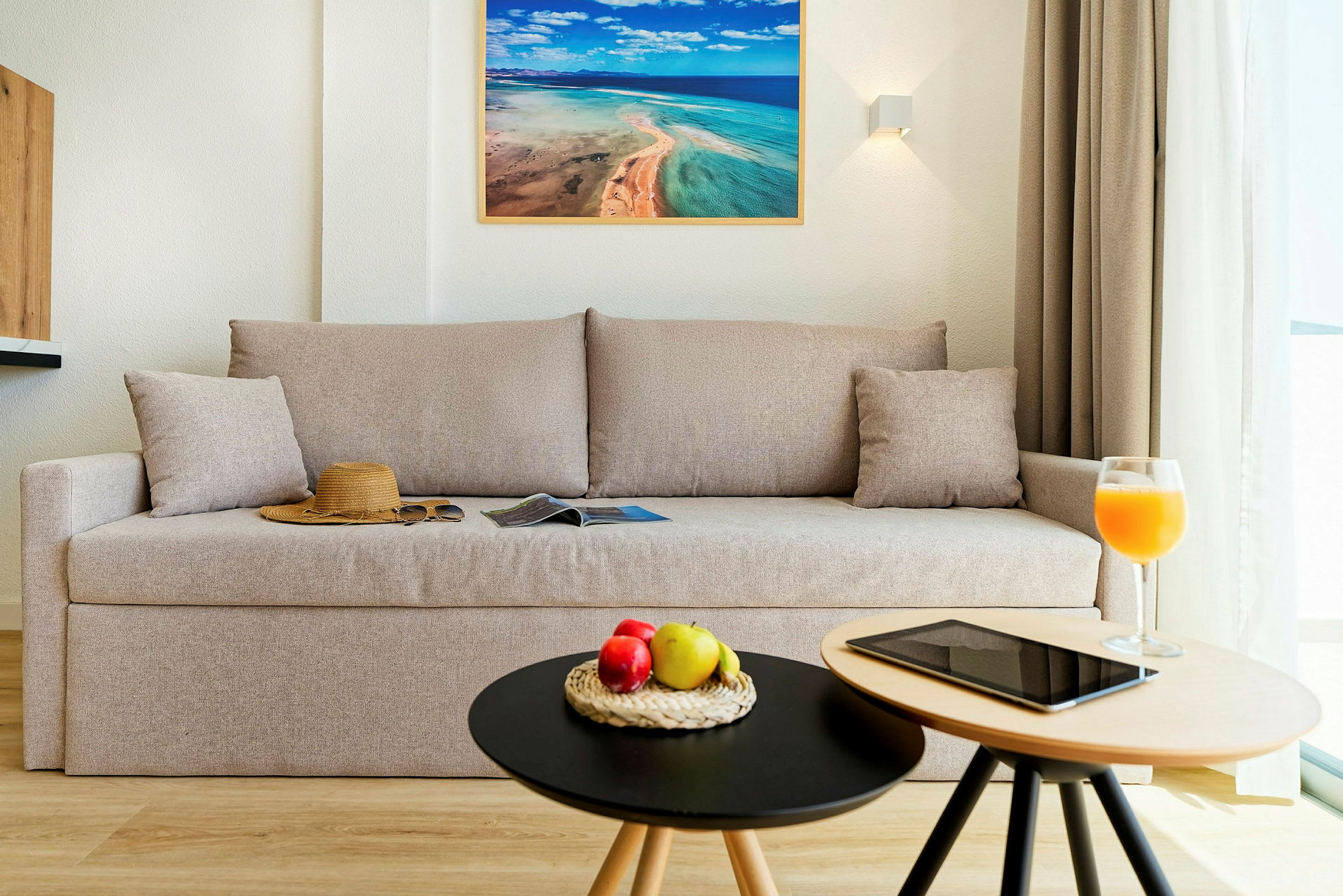 En stue med en beige sofa, en hatt og solbriller på sofaen, og to små bord med frukt, et nettbrett og et glass juice. (AI-generert tekst)