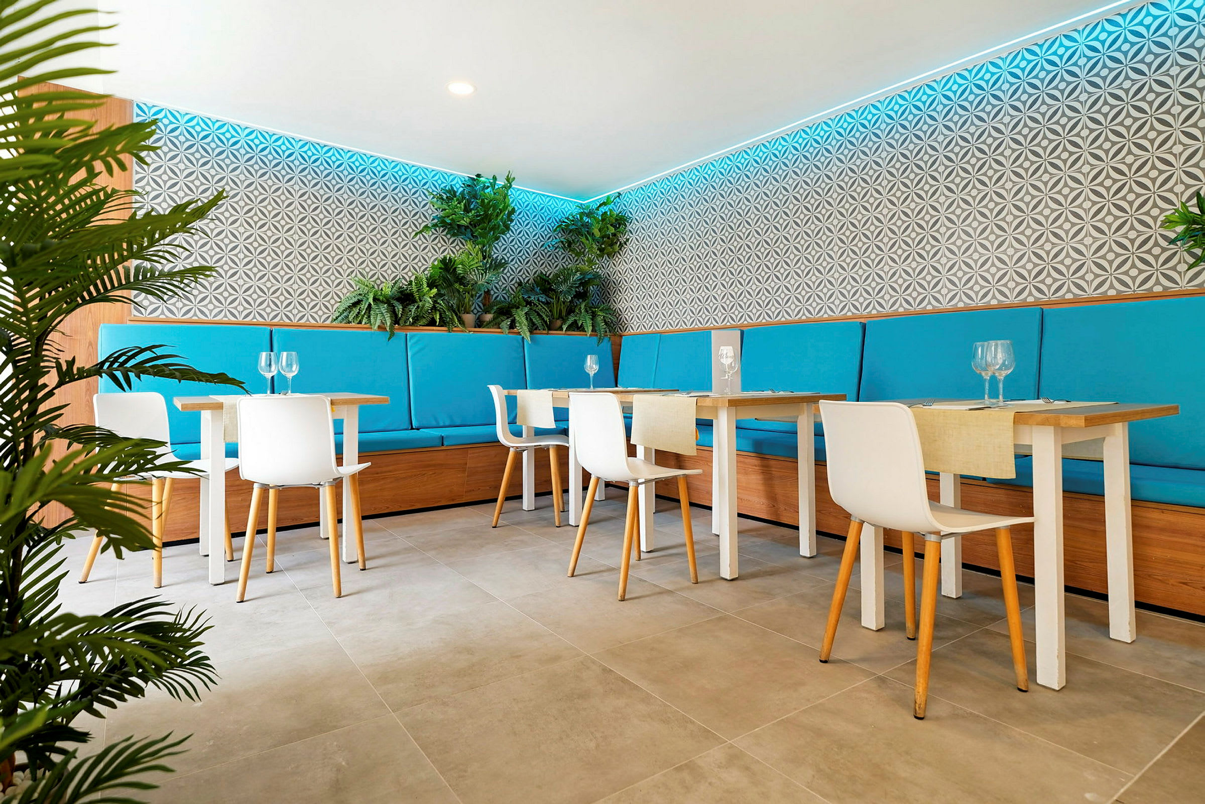 En moderne restaurant med lyse møbler, turkise sitteputer og planter som dekorasjon langs veggene. (AI-generert tekst)