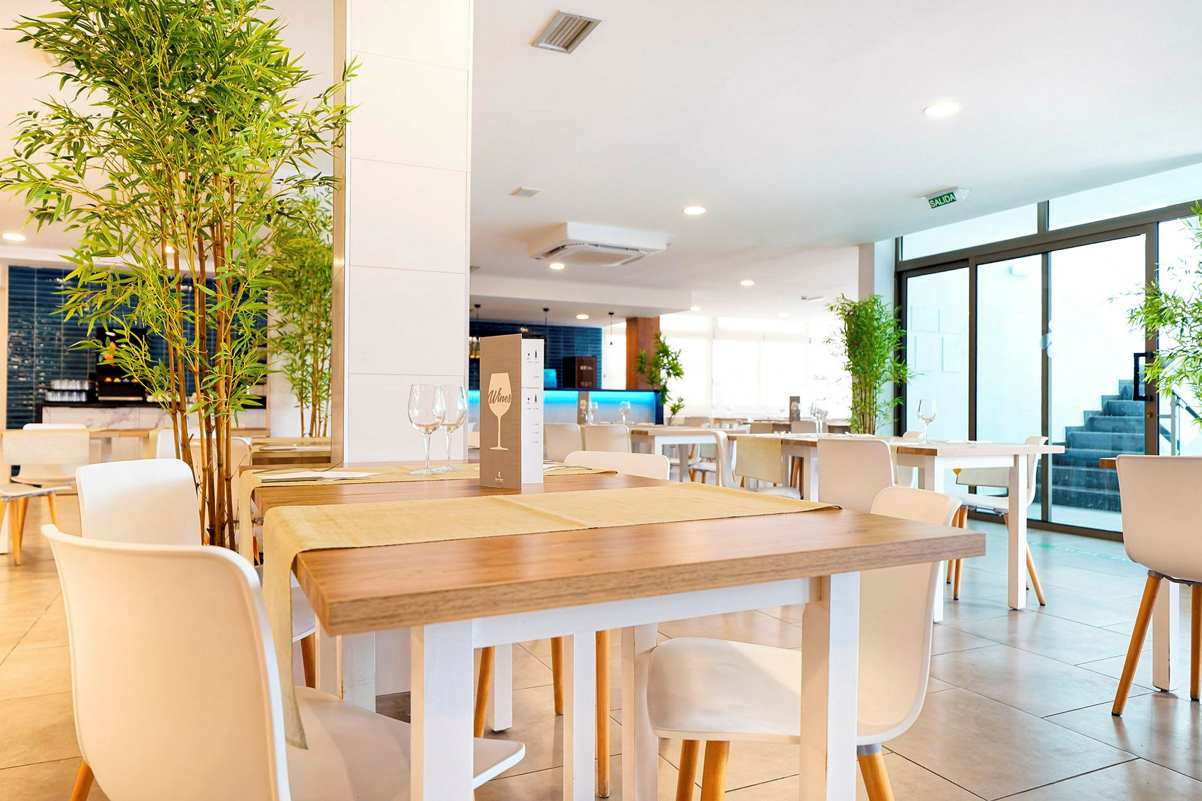 Et moderne og lyst innredet restaurantområde med trebord og hvite stoler samt dekorative planter plassert i hjørnene. (AI-generert tekst)