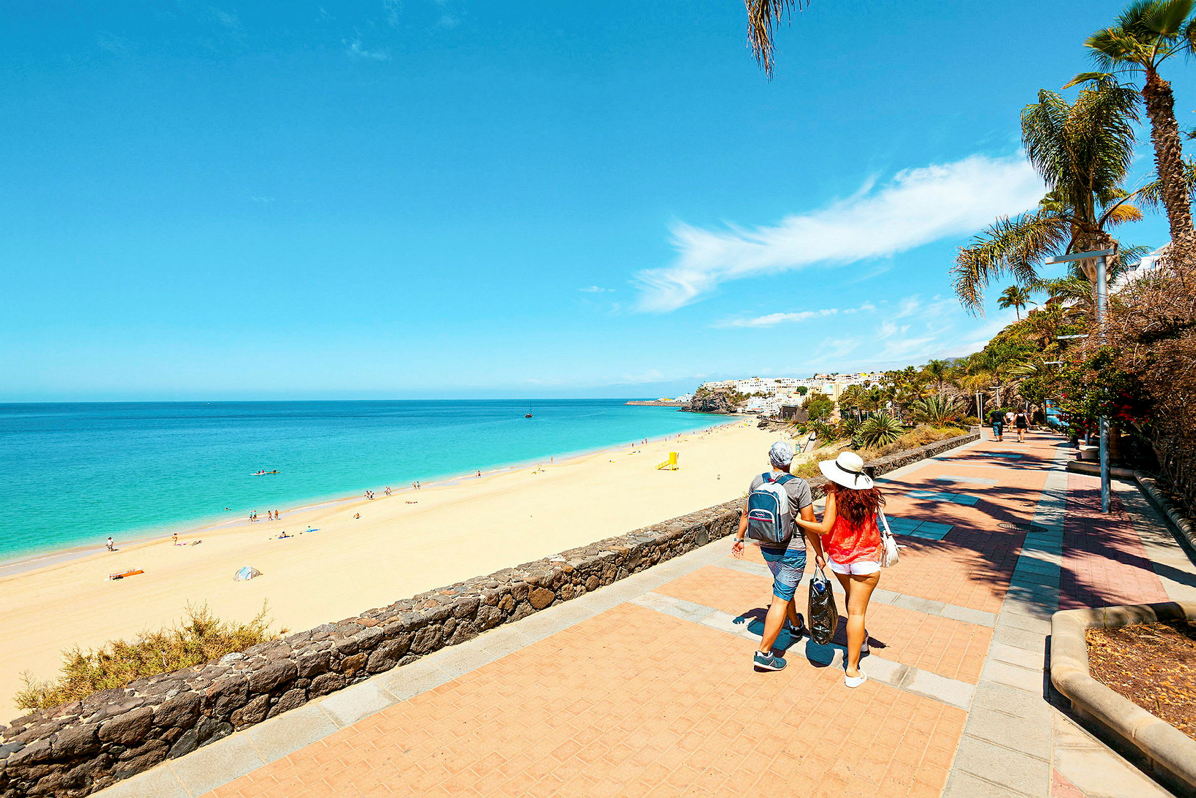 En strand med gyllen sand og klart blått hav, omgitt av en promenade med palmer og annen vegetasjon. En person går langs promenaden. (AI-generert tekst)