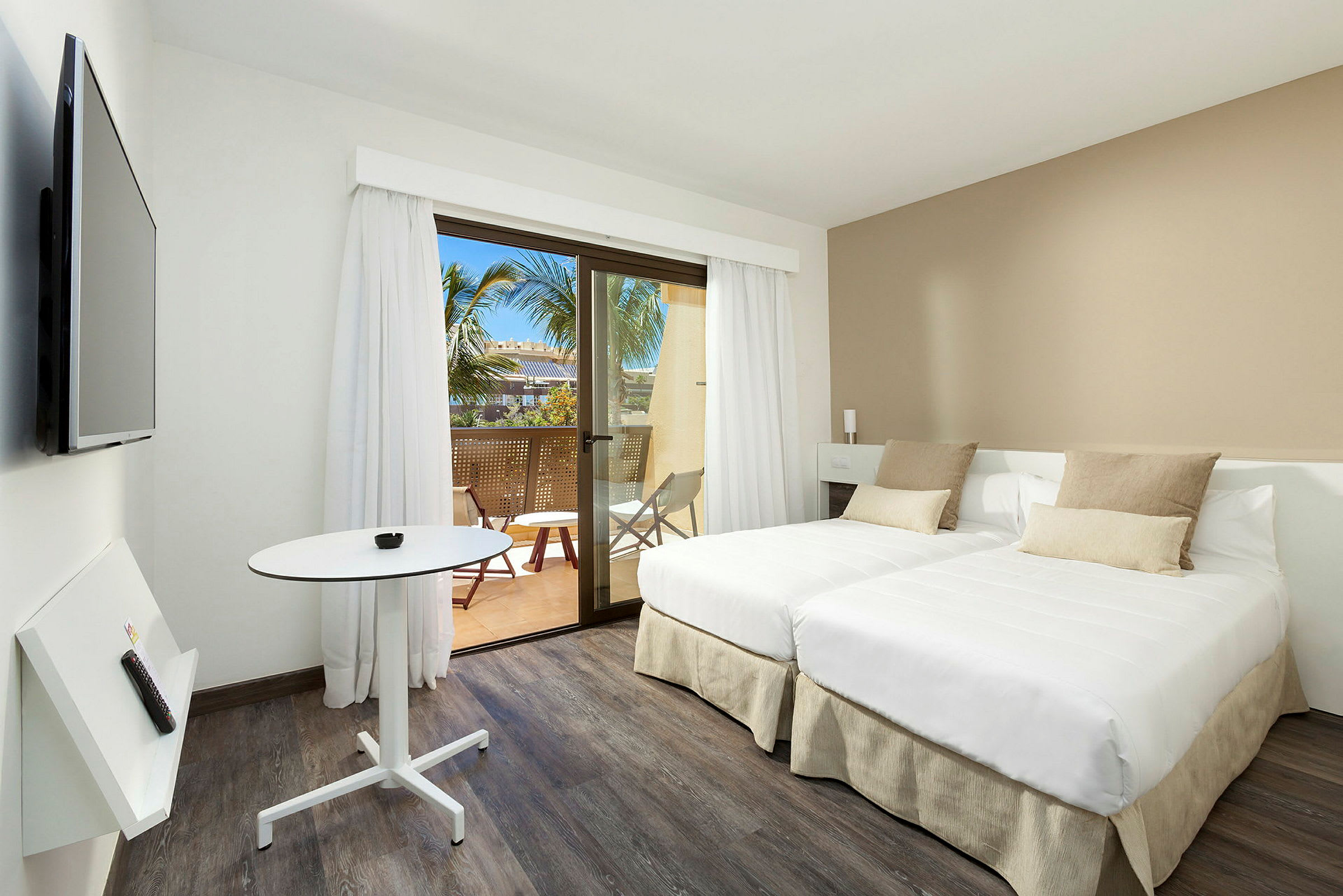 Et moderne hotellrom med to enkeltsenger, et lite bord og en veggmontert TV. Rommet har lyse farger og et vindu som leder ut til en balkong med utsikt mot palmer. (AI-generert tekst)