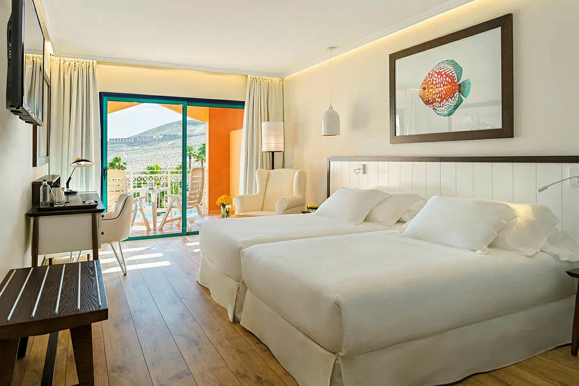 Et lyst og moderne hotellrom med to senger, en sittegruppe og et skrivebord. En balkong med utsikt over landskapet er synlig i bakgrunnen. (AI-generert tekst)