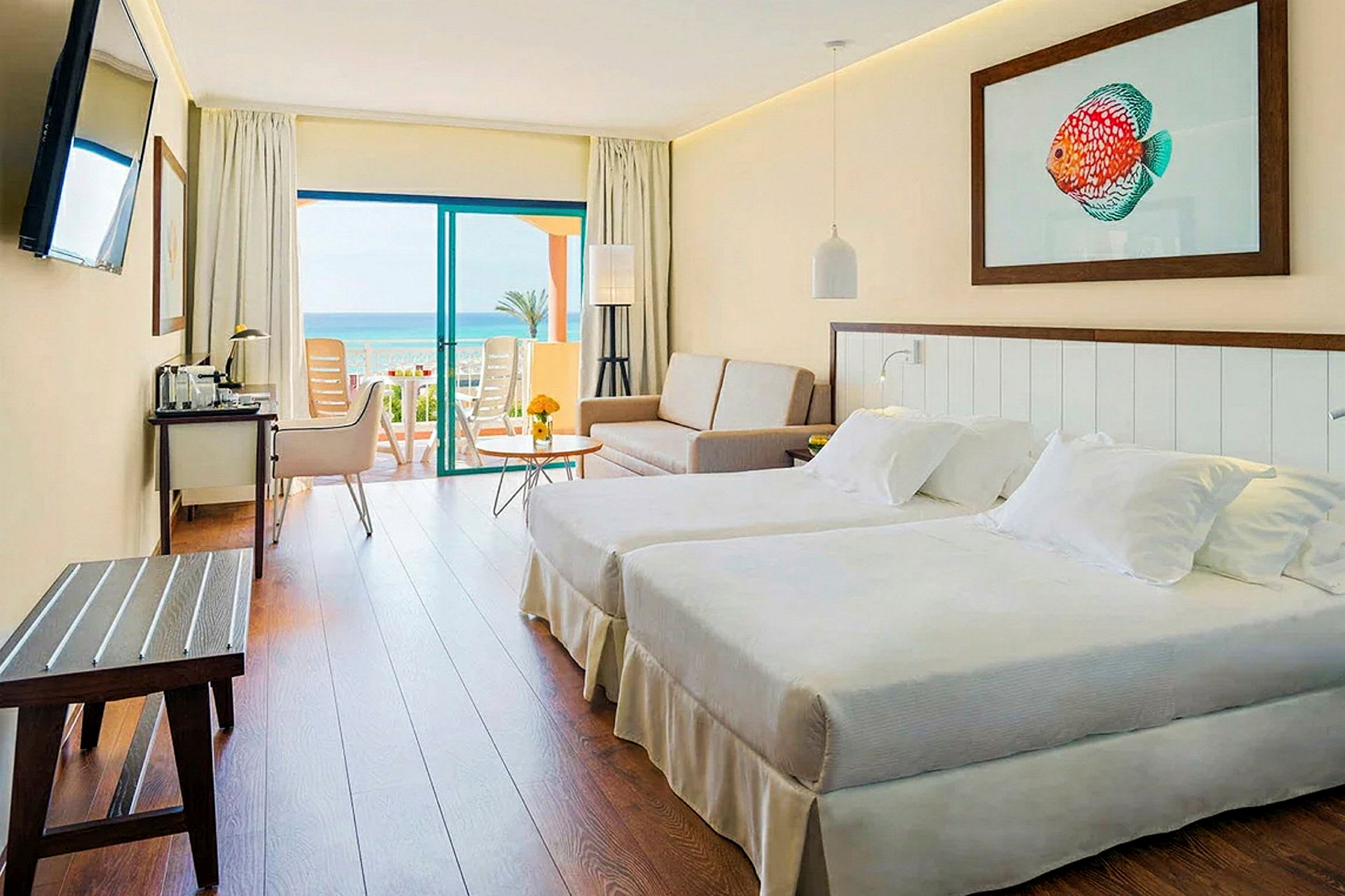 Et hotellrom med to senger, en sofa og et bord. Rommet har en balkongdør som leder til en balkong med utsikt over havet. (AI-generert tekst)