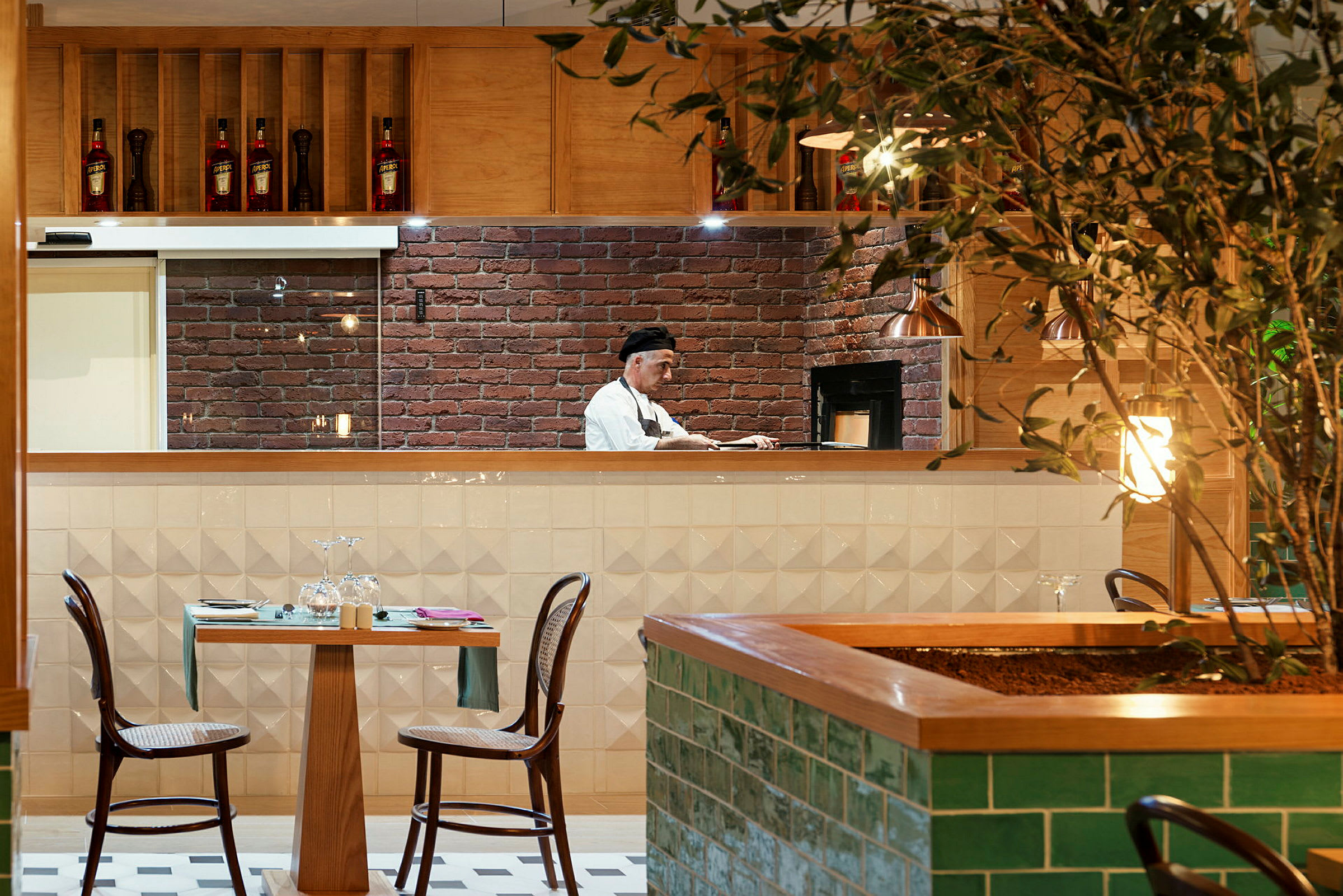 En restaurant med et åpent kjøkken hvor en person arbeider. Et bord med to stoler står foran kjøkkenet, og en plante er synlig i forgrunnen. (AI-generert tekst)