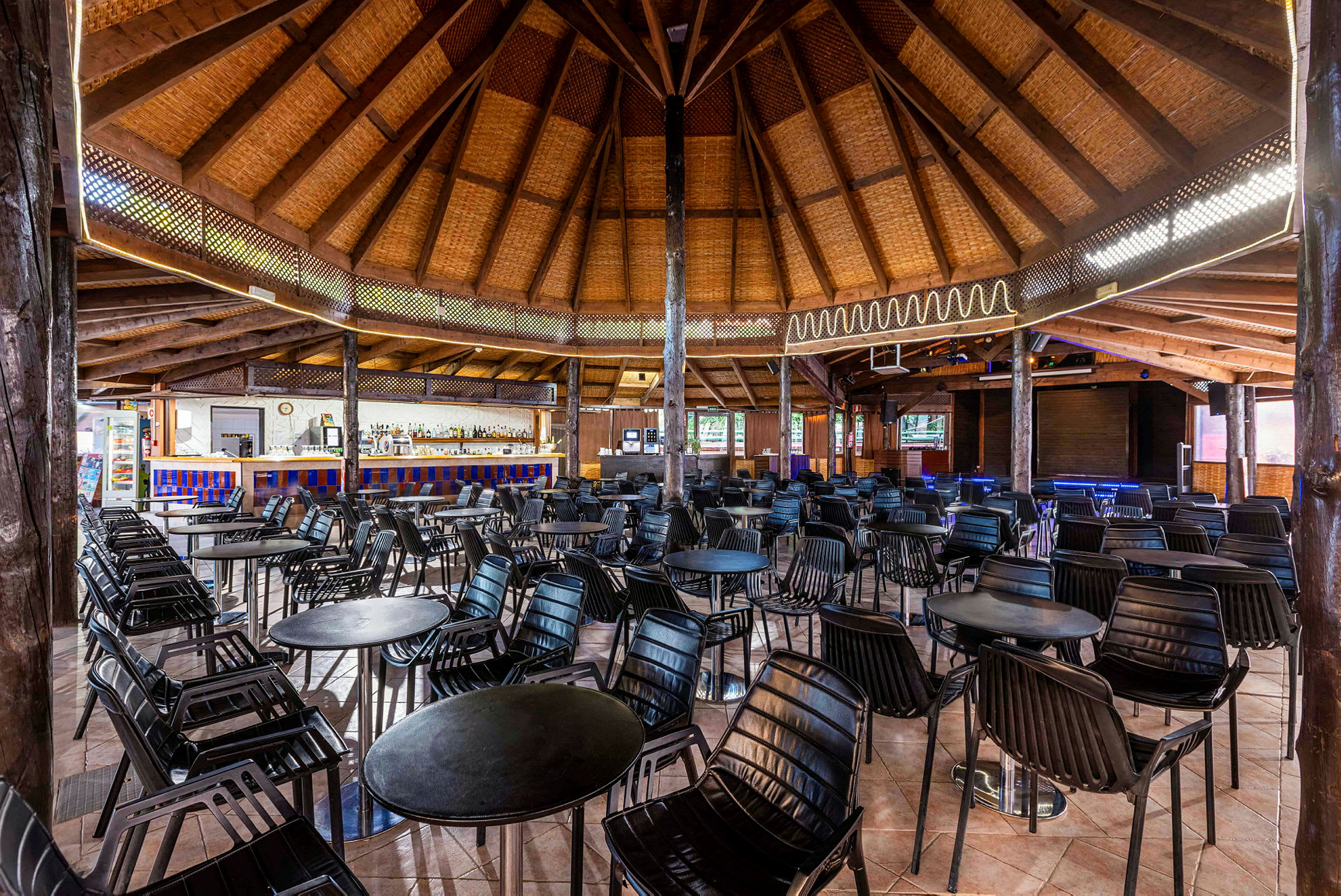 Et rundt trehus med stråtak inneholder en innredet hall med bord og stoler og en bar i bakgrunnen. (AI-generert tekst)