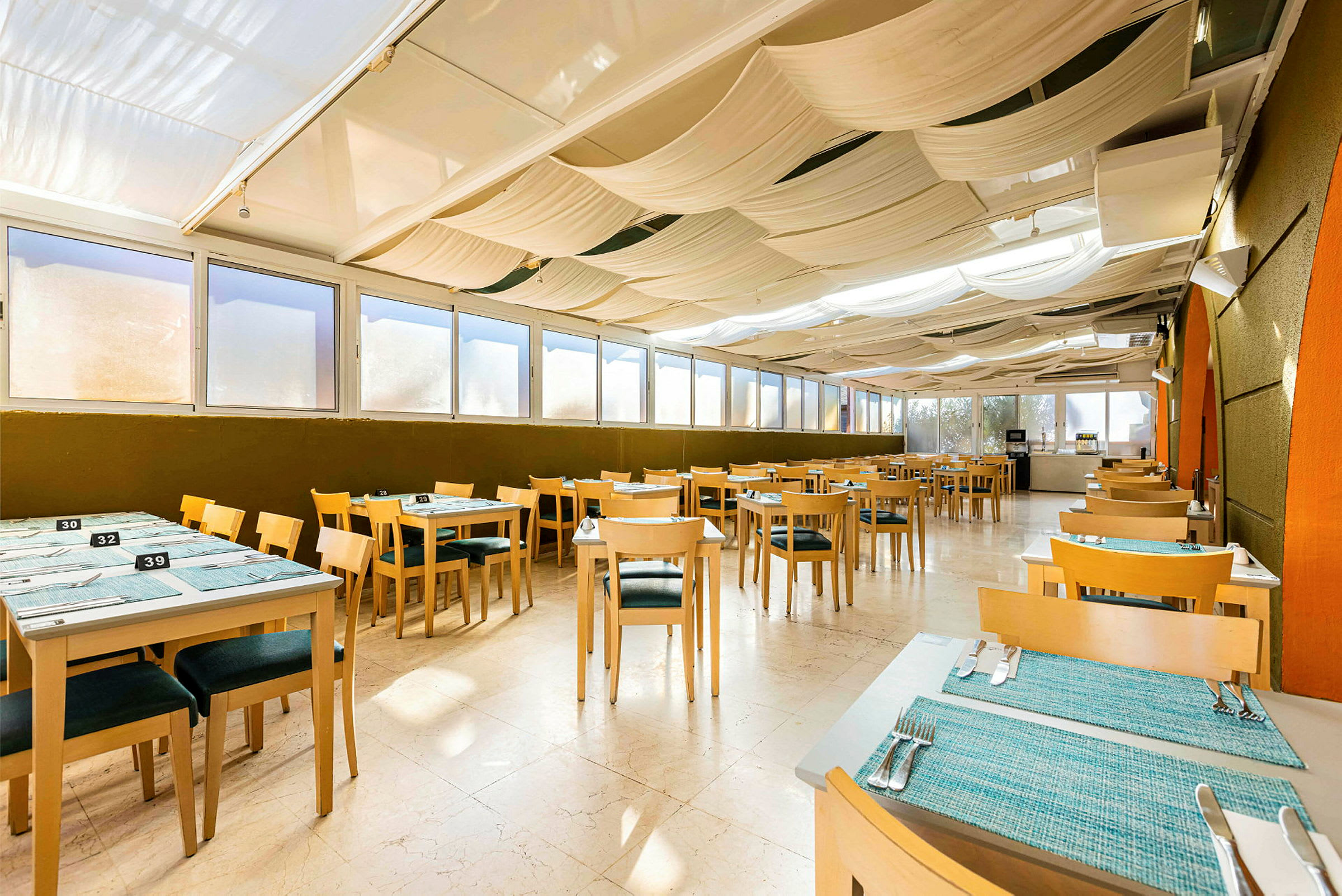 Bilde av en innendørs restaurant med trebord og stoler, blå bordmatter og dagslys fra store vinduer. (AI-generert tekst)
