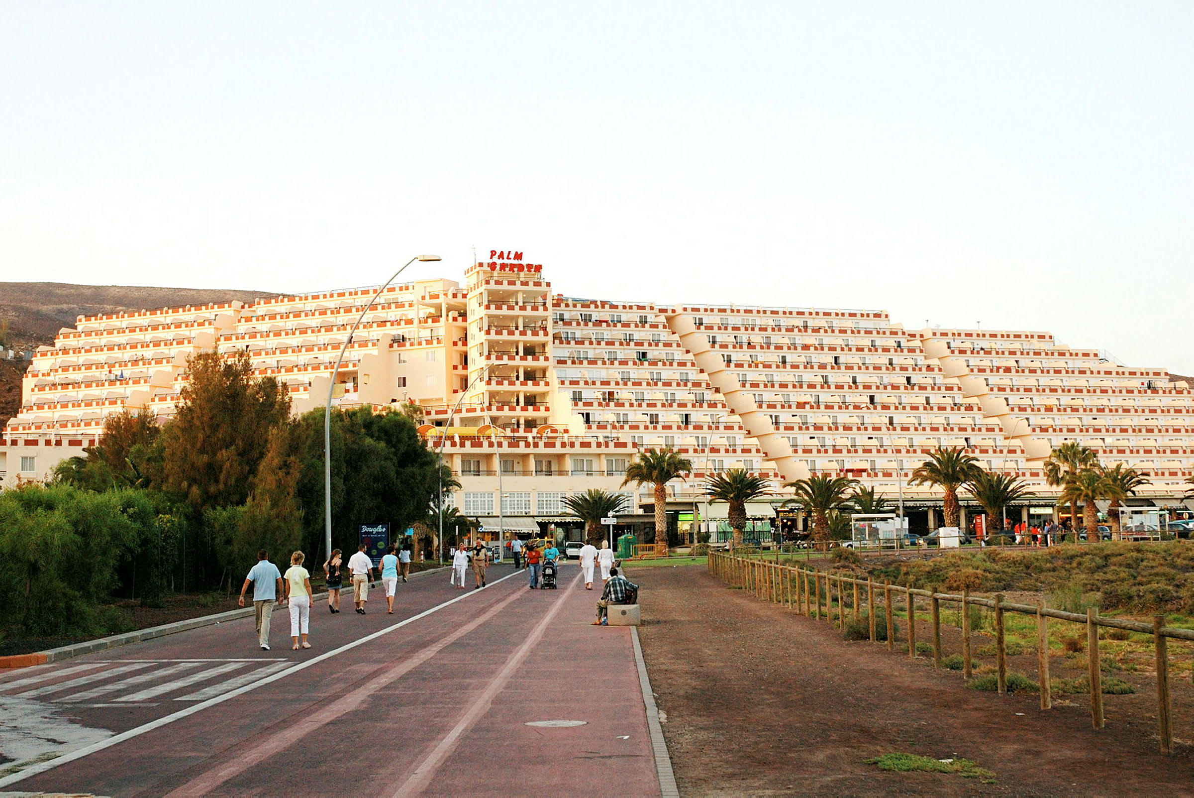 En stor bygning i pyramideform med flere etasjer ligger foran en gangvei med folk som går og nyter miljøet. Trær og vegetasjon er synlig i forgrunnen. (AI-generert tekst)