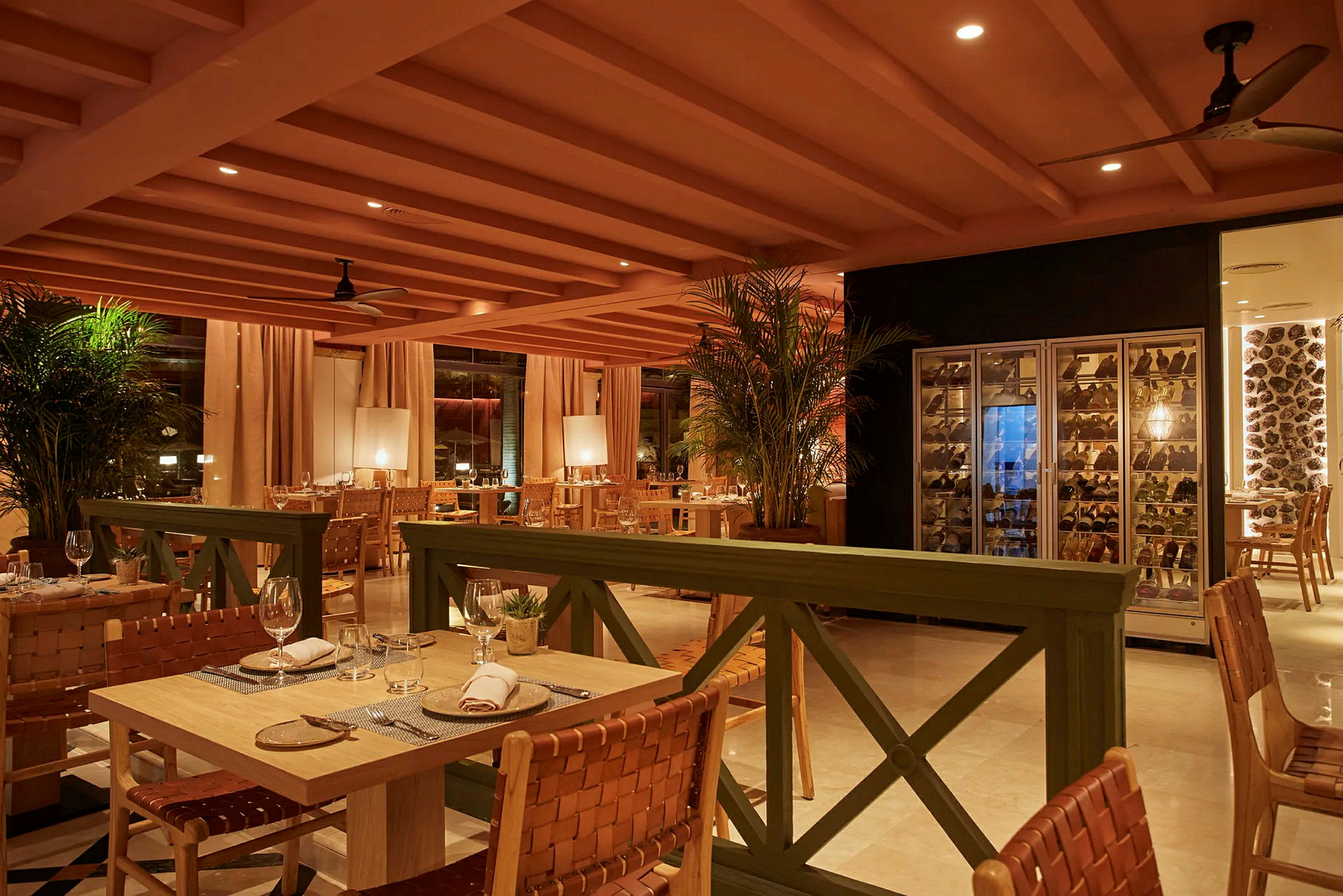 Bilde av en elegant restaurant med bord og stoler, vinlagringsskap til høyre og varm belysning. (AI-generert tekst)