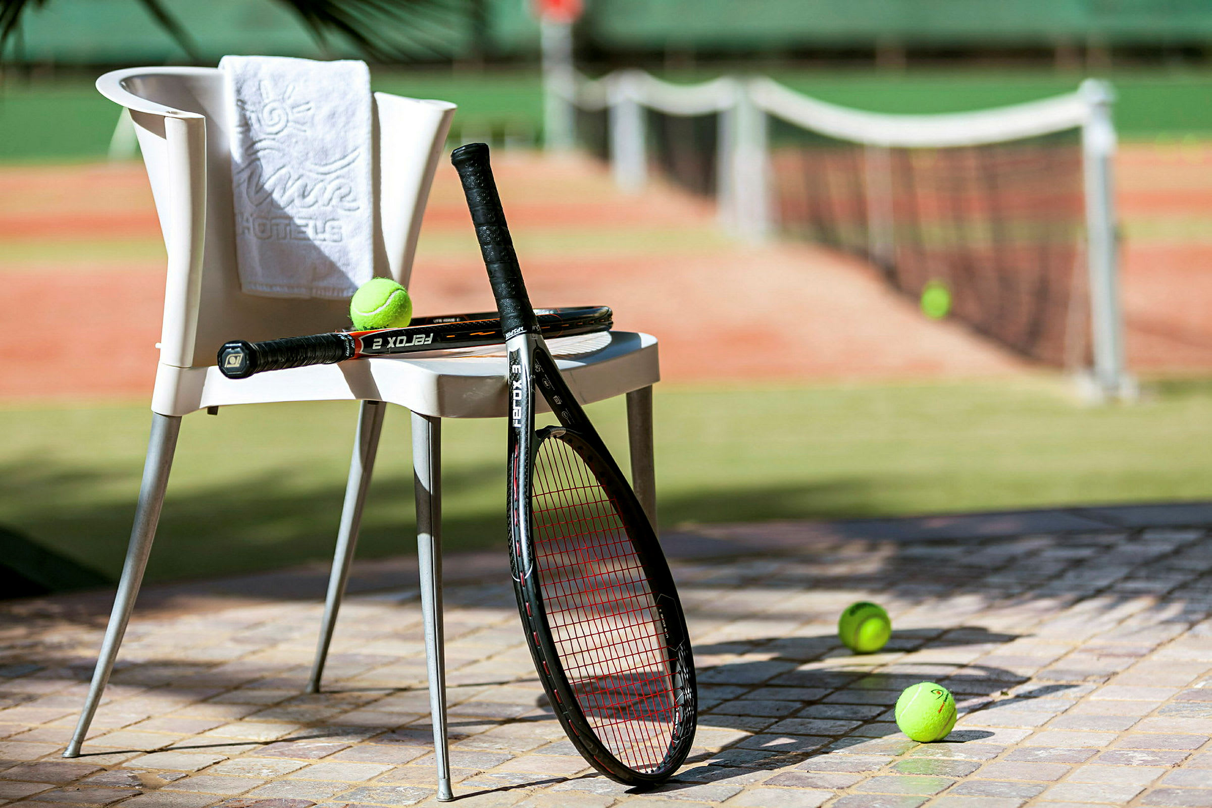 En stol med et håndkle og to tennisracketer plassert ved siden av en tennisbane, med tennisballer på bakken. (AI-generert tekst)