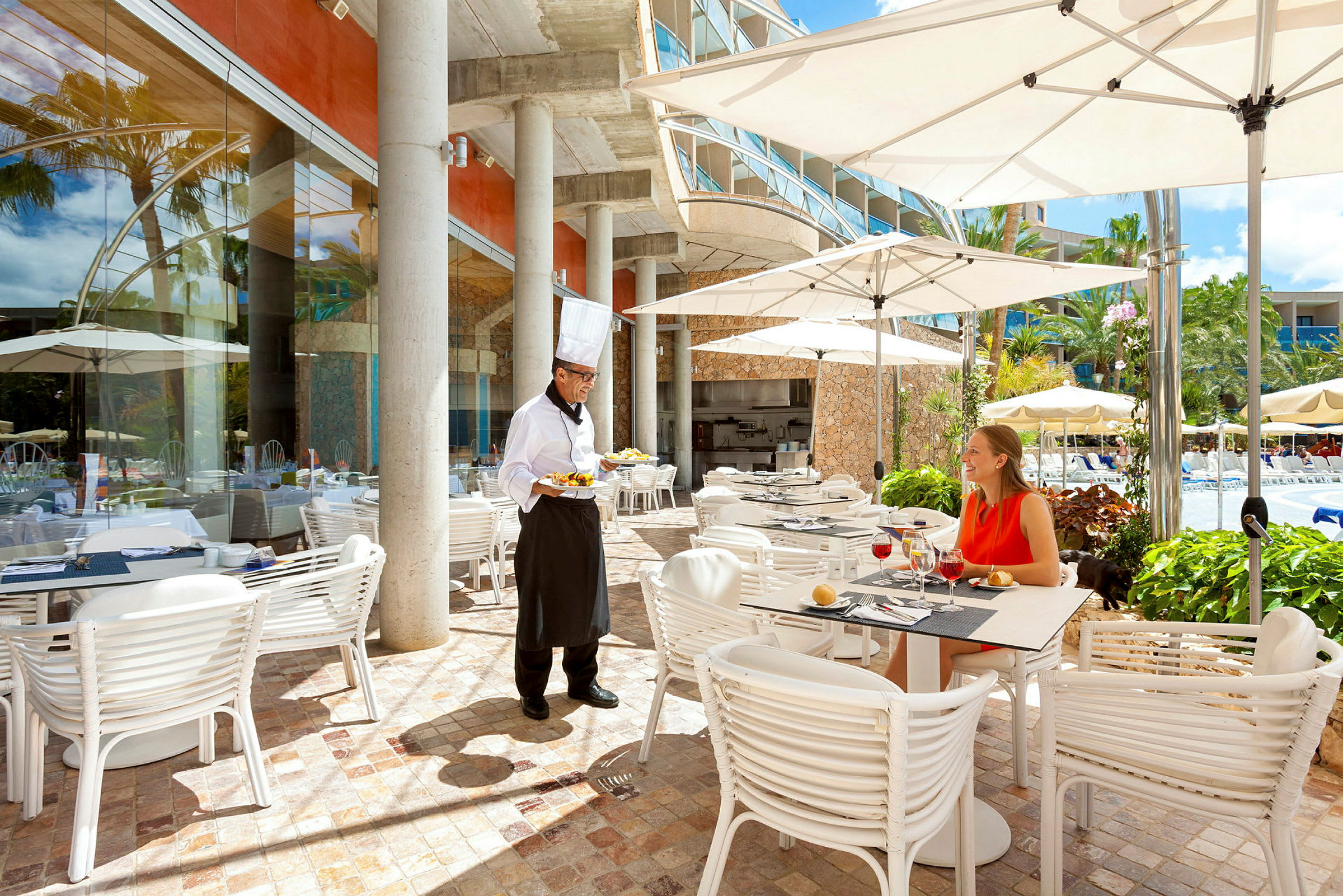 En restaurantterrasse med bord, stoler og parasoller ved siden av et basseng. En kelner serverer mat til en gjest. (AI-generert tekst)