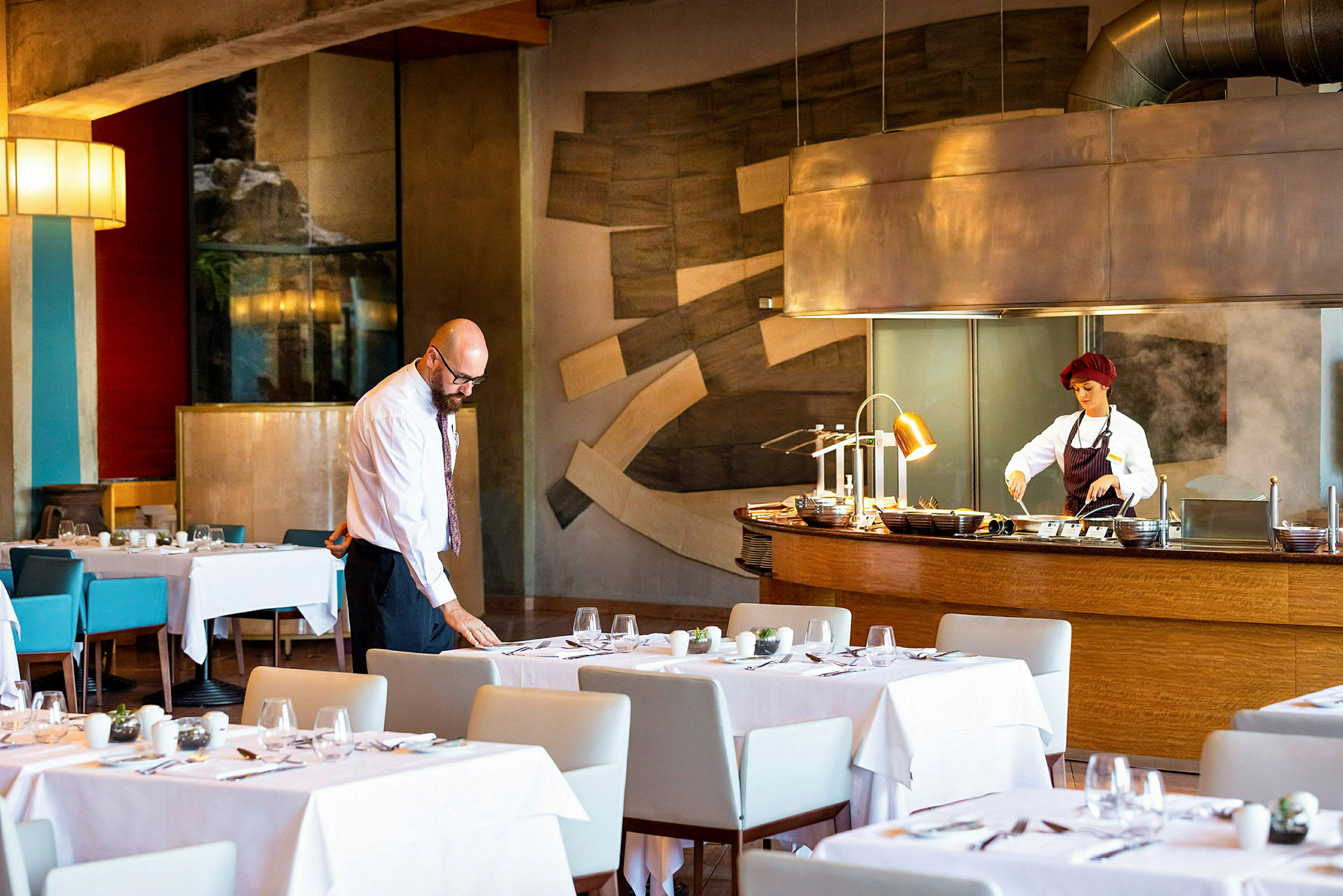 Restaurantinteriør med dekket bord og en matlagingsstasjon med en person i arbeidsklær. (AI-generert tekst)