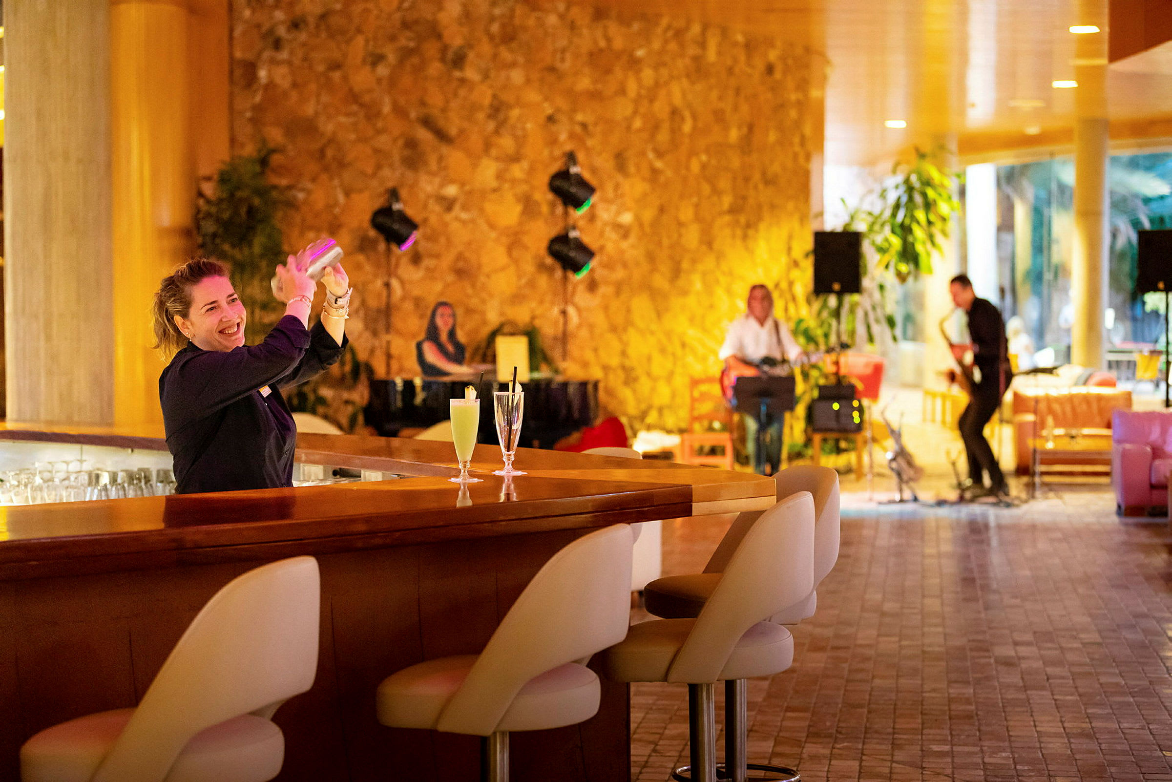 En bartender lager en drink ved bardisken. I bakgrunnen kan man se musikere spille instrumenter i et varmt og stilfullt miljø. (AI-generert tekst)