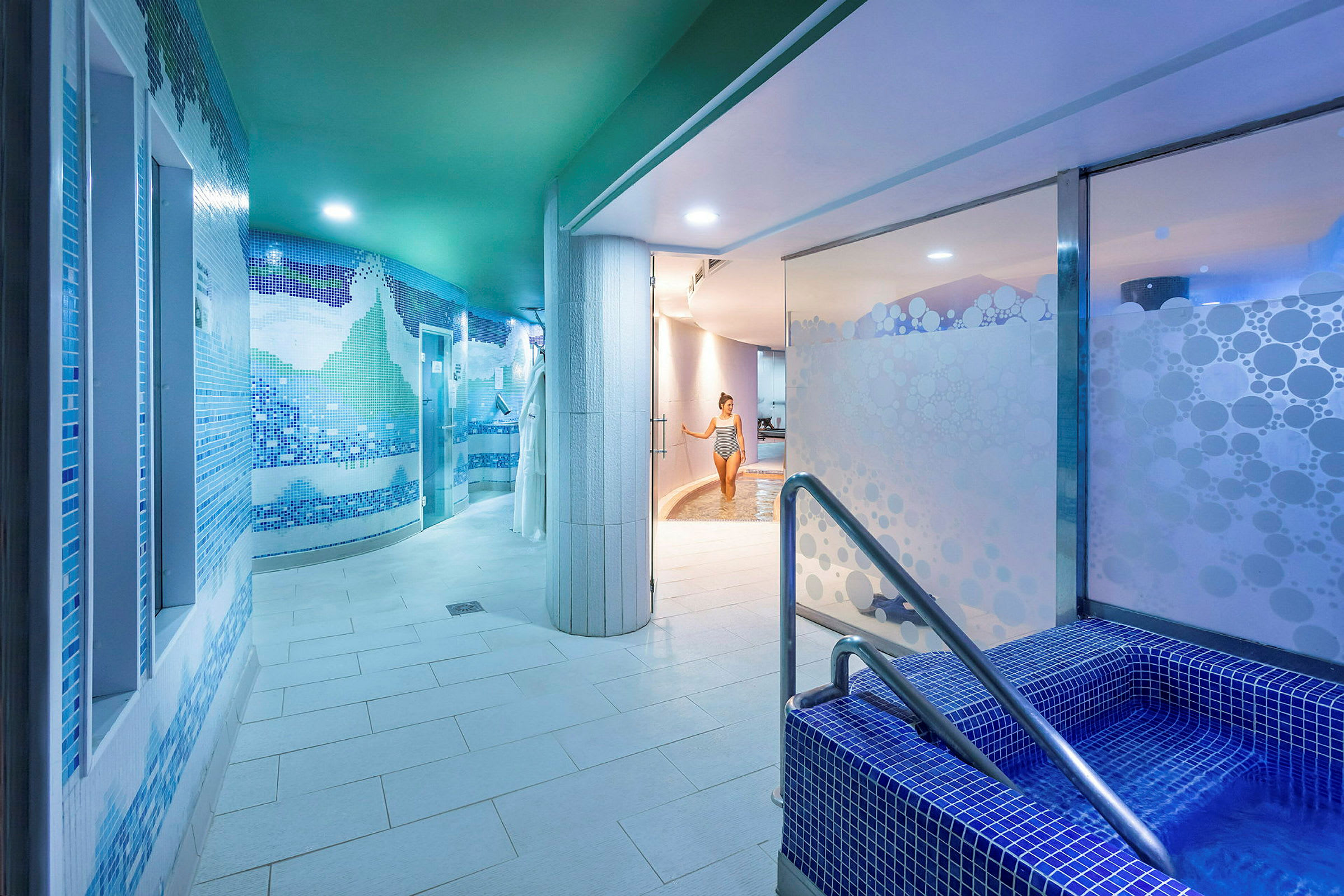 Et innendørs spa med blå mosaikkvegger, et mindre basseng med trapper i forgrunnen og en person som går mot et større basseng i bakgrunnen. (AI-generert tekst)