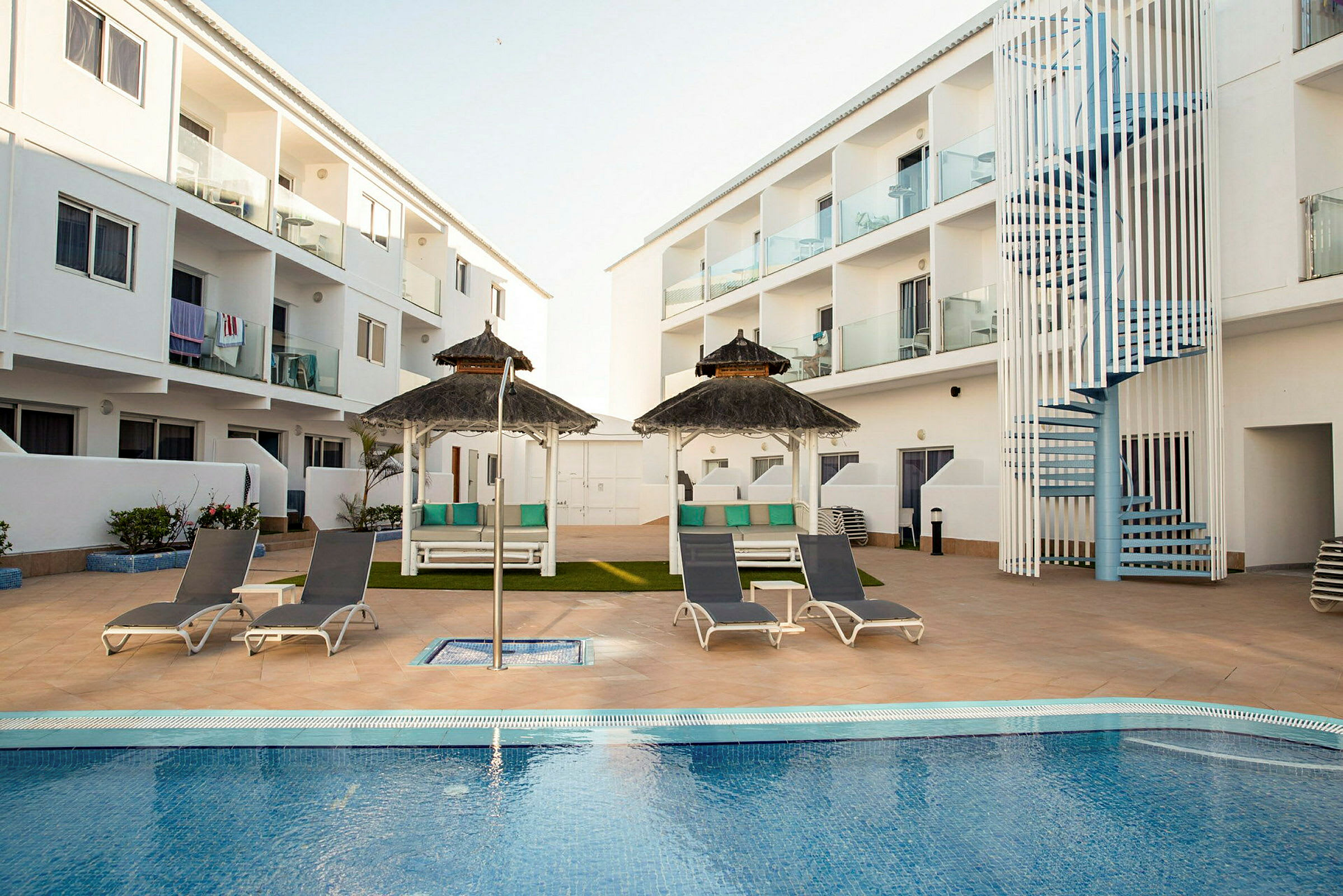 Et hotellområde med et svømmebasseng omgitt av solsenger og parasoller. I bakgrunnen sees hvite bygninger med balkonger. (AI-generert tekst)