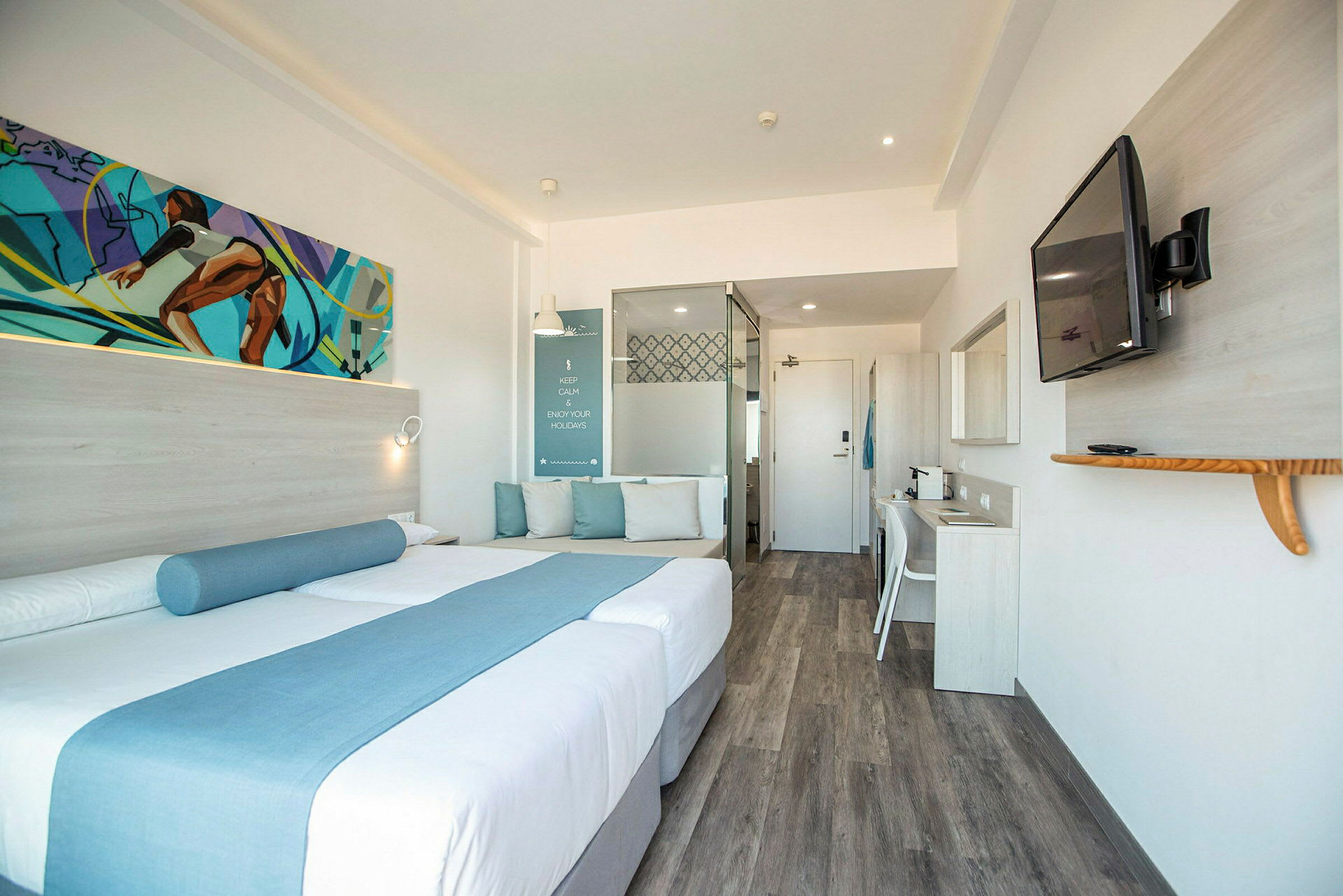 Et moderne hotellrom med en dobbeltseng dekorert med blå tekstiler, en sitteplass med puter, et skrivebord og en veggmontert TV. (AI-generert tekst)