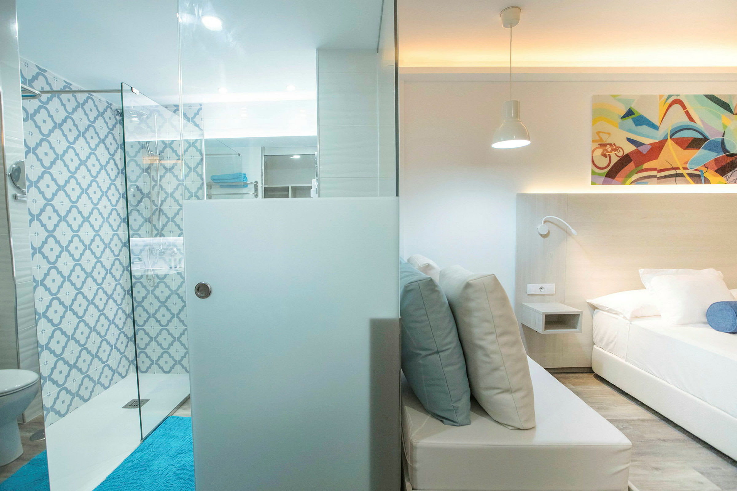 Et hotellrom med sitteområde og seng, adskilt av en glassvegg. Badet har blått mønstret fliser og dusj. (AI-generert tekst)