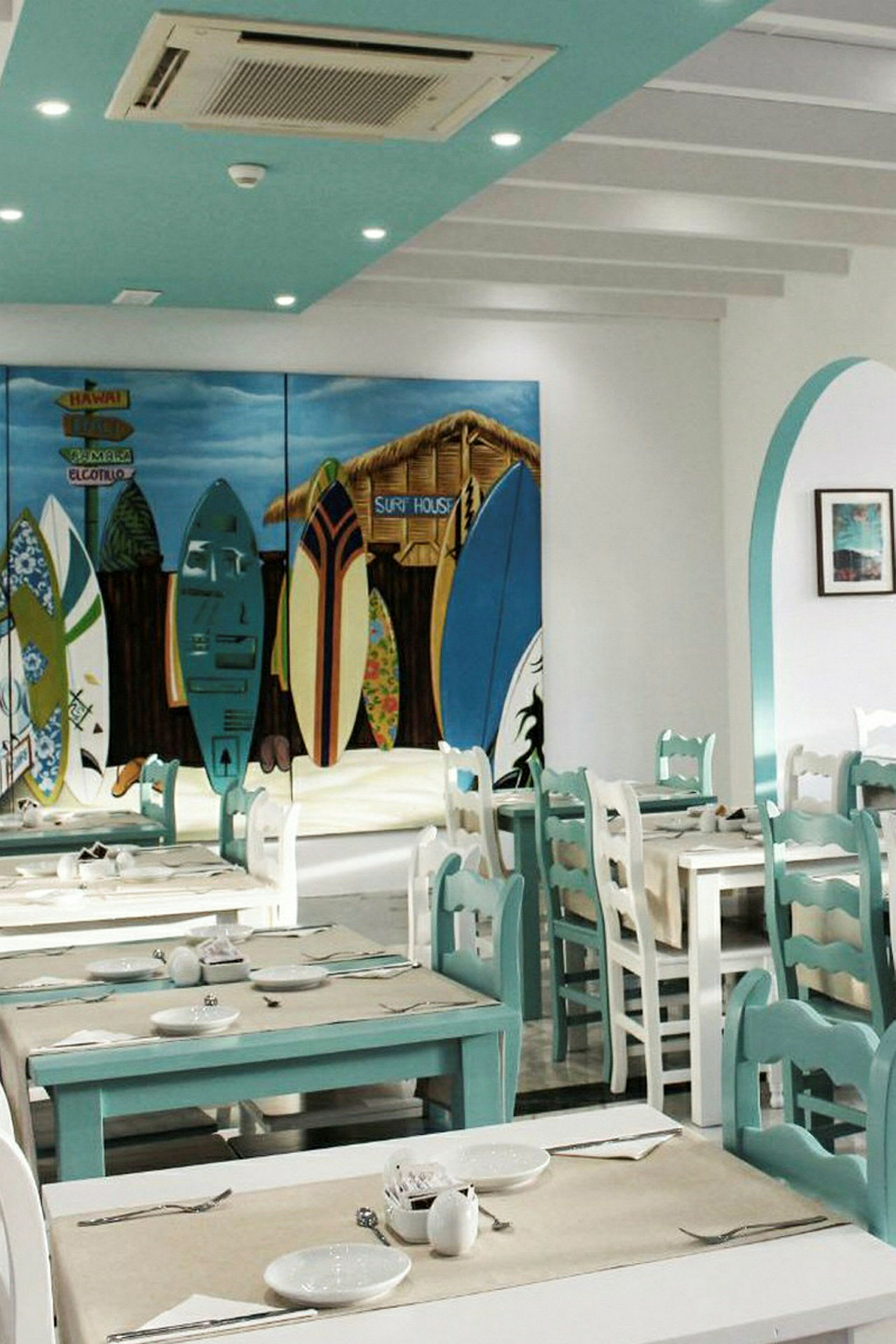 En interiør av en restaurant med turkise og hvite bord og stoler. Et fargerikt veggmaleri viser surfebrett og en hytte. (AI-generert tekst)