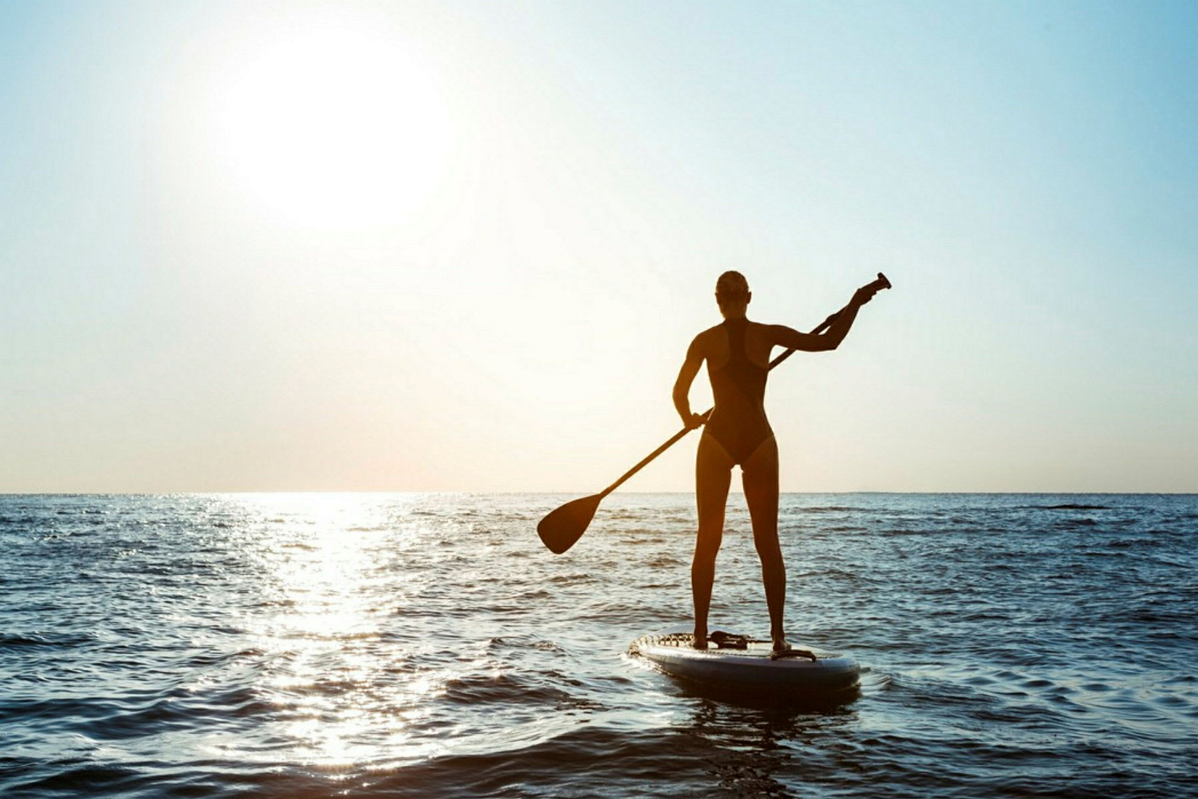 En person padler stående på et paddleboard på havet i motlys med solen i bakgrunnen. (AI-generert tekst)
