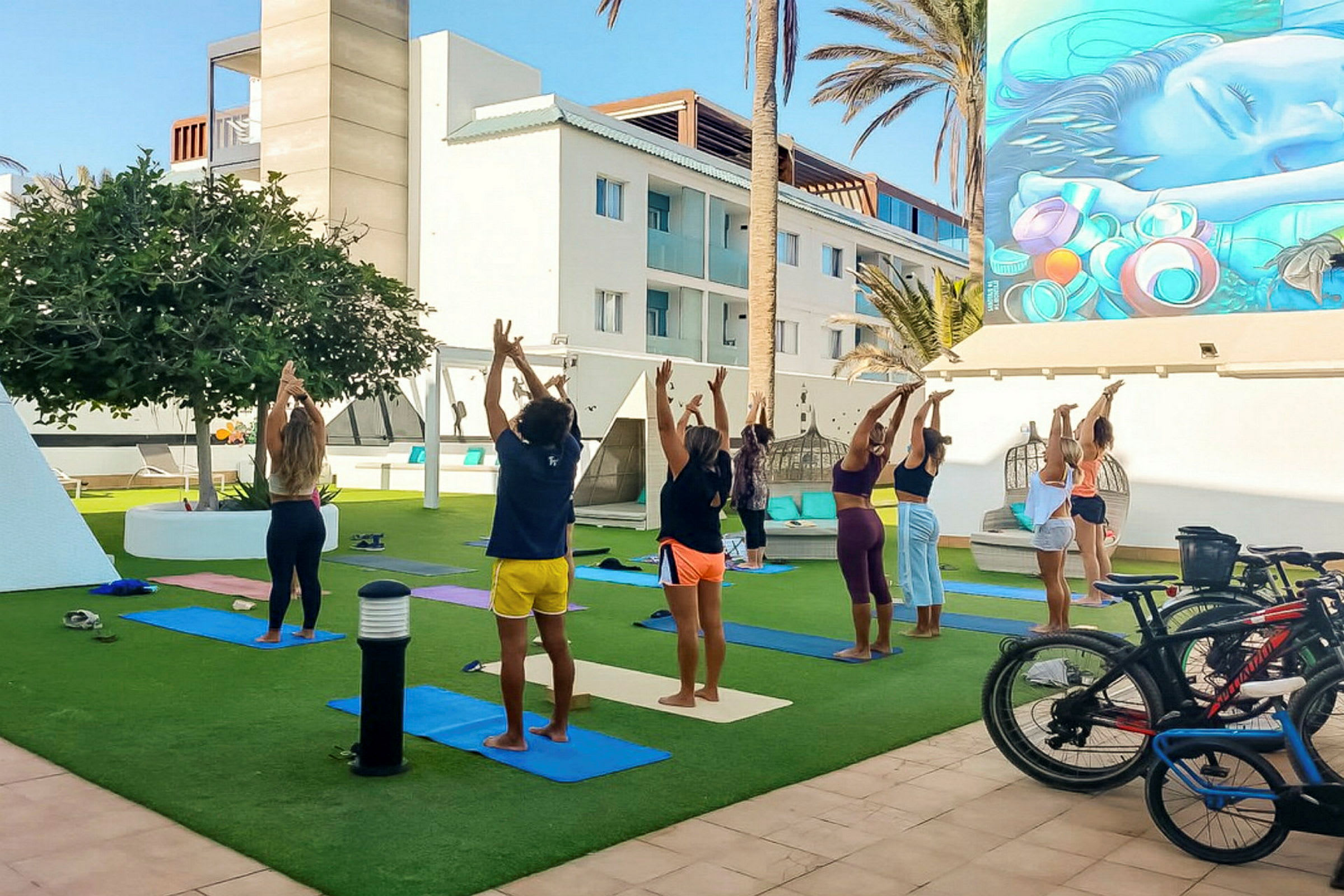 En gruppe mennesker utfører yoga utendørs på gress med matter, omgitt av en moderne bygning og palmer. (AI-generert tekst)