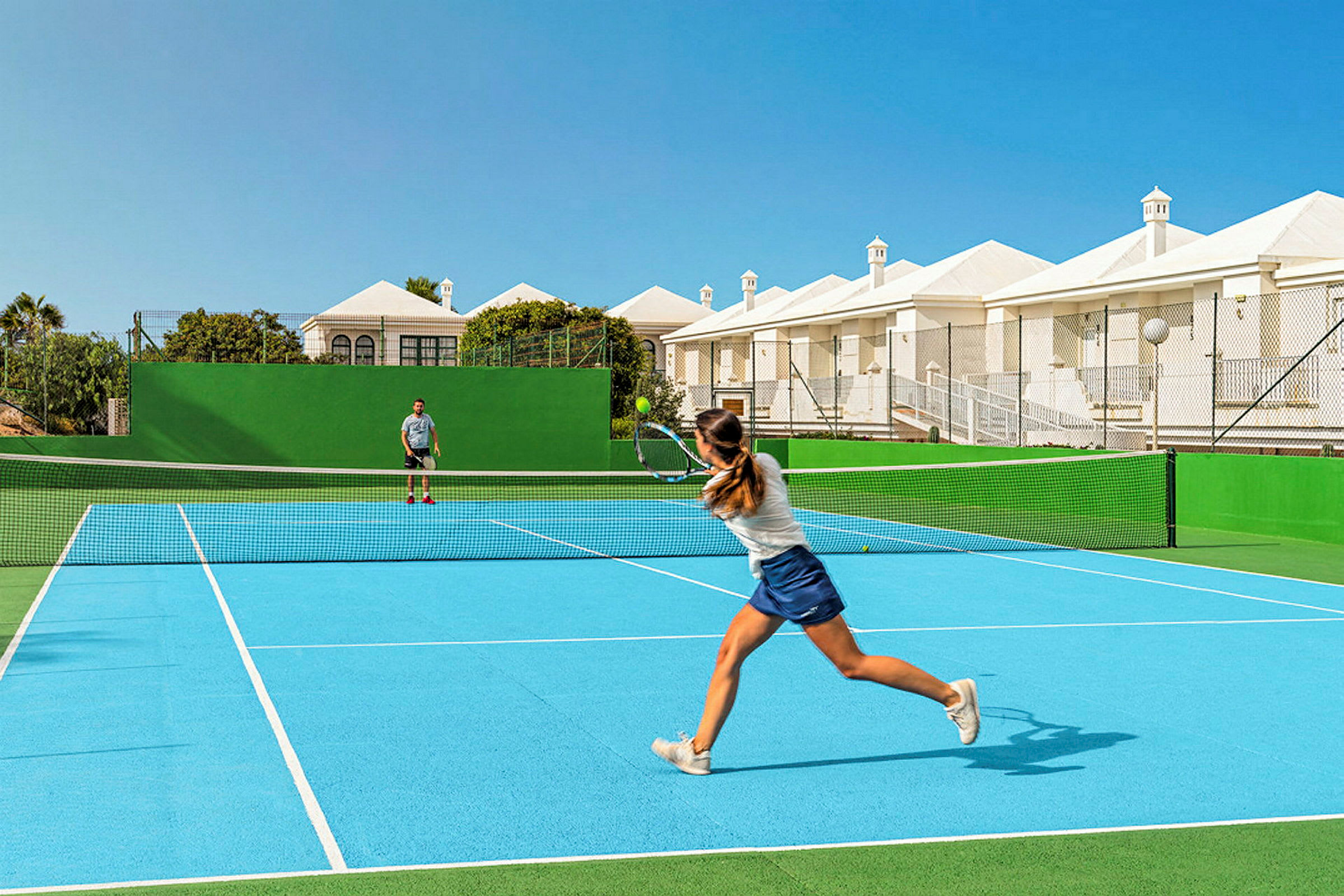 To personer spiller tennis på en utendørs tennisbane med blått underlag og grønne omgivelser. Bak banen sees hvite bygninger og blå himmel. (AI-generert tekst)