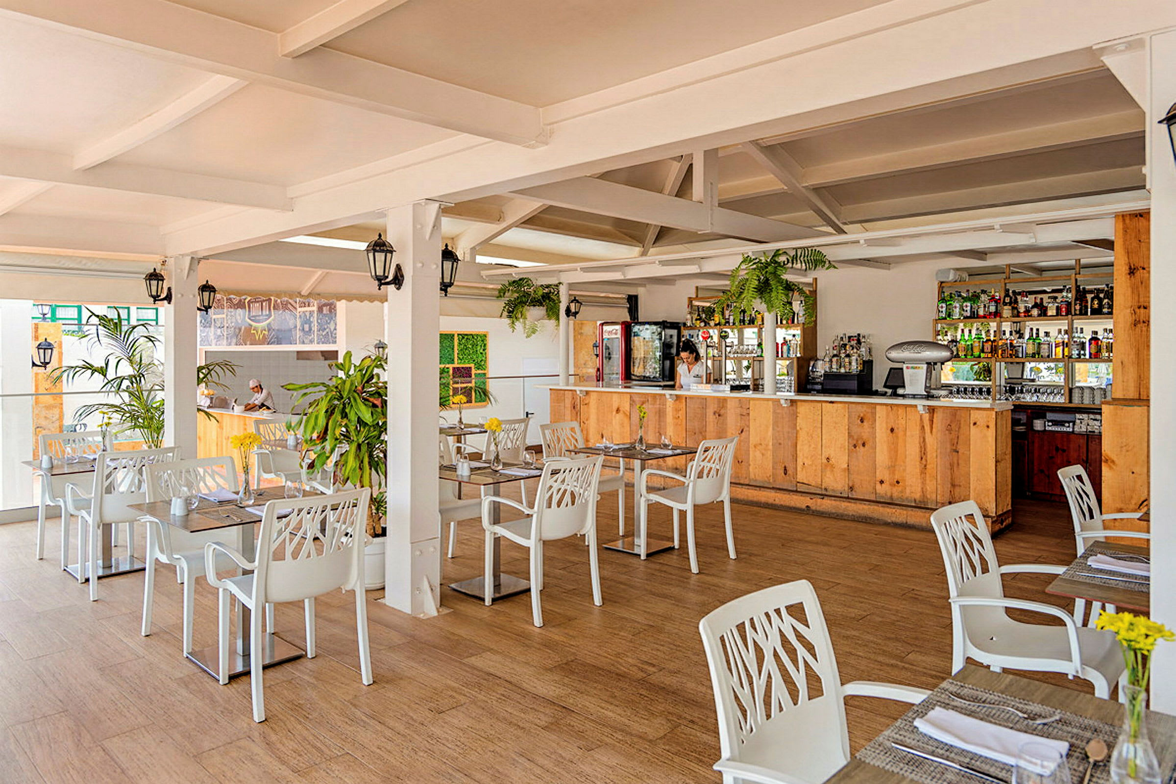 En lys restaurant med hvite bord og stoler samt en bardisk i tre. (AI-generert tekst)