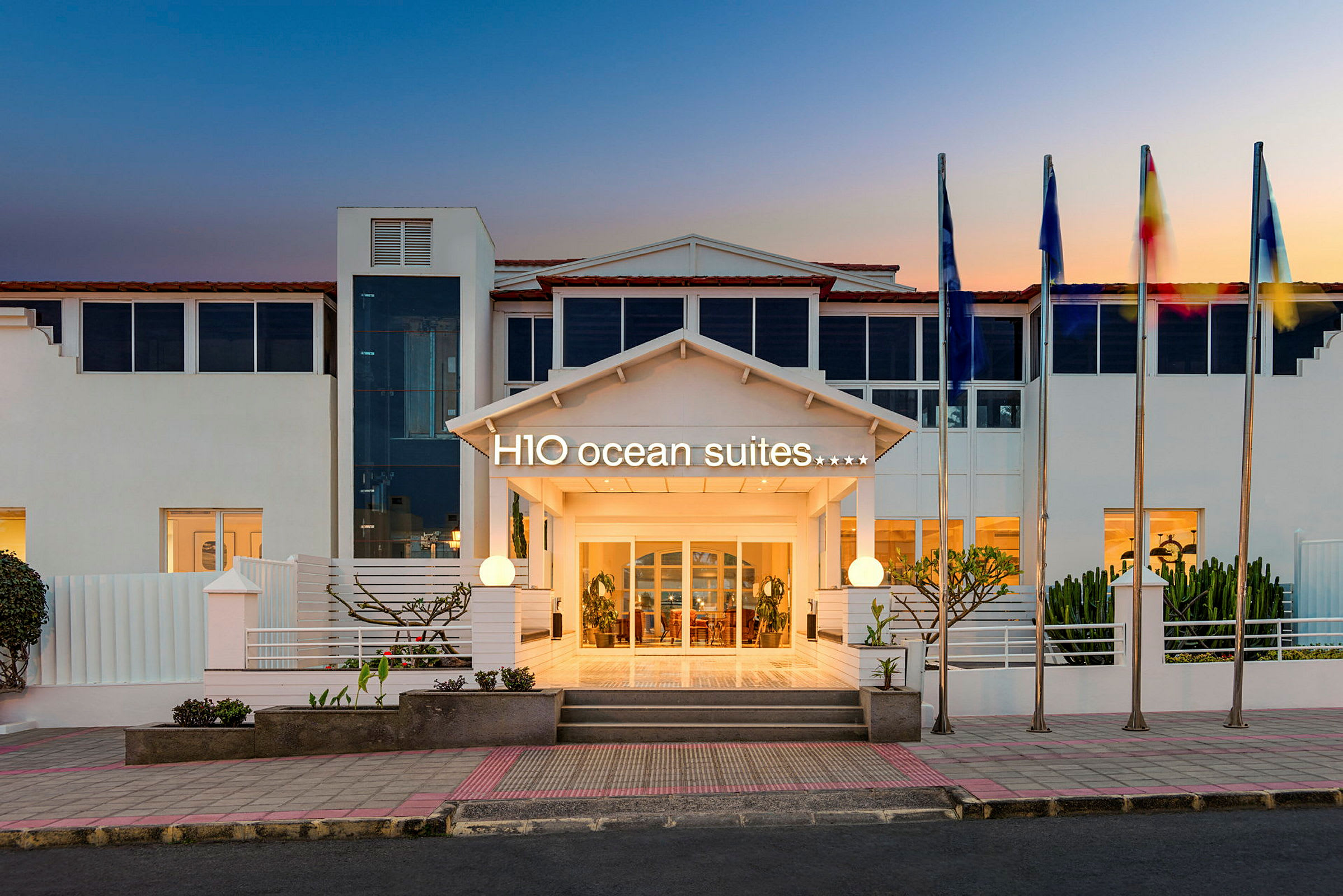 Hotellinngang med skiltet H10 Ocean Suites og flaggstenger ved solnedgang. (AI-generert tekst)