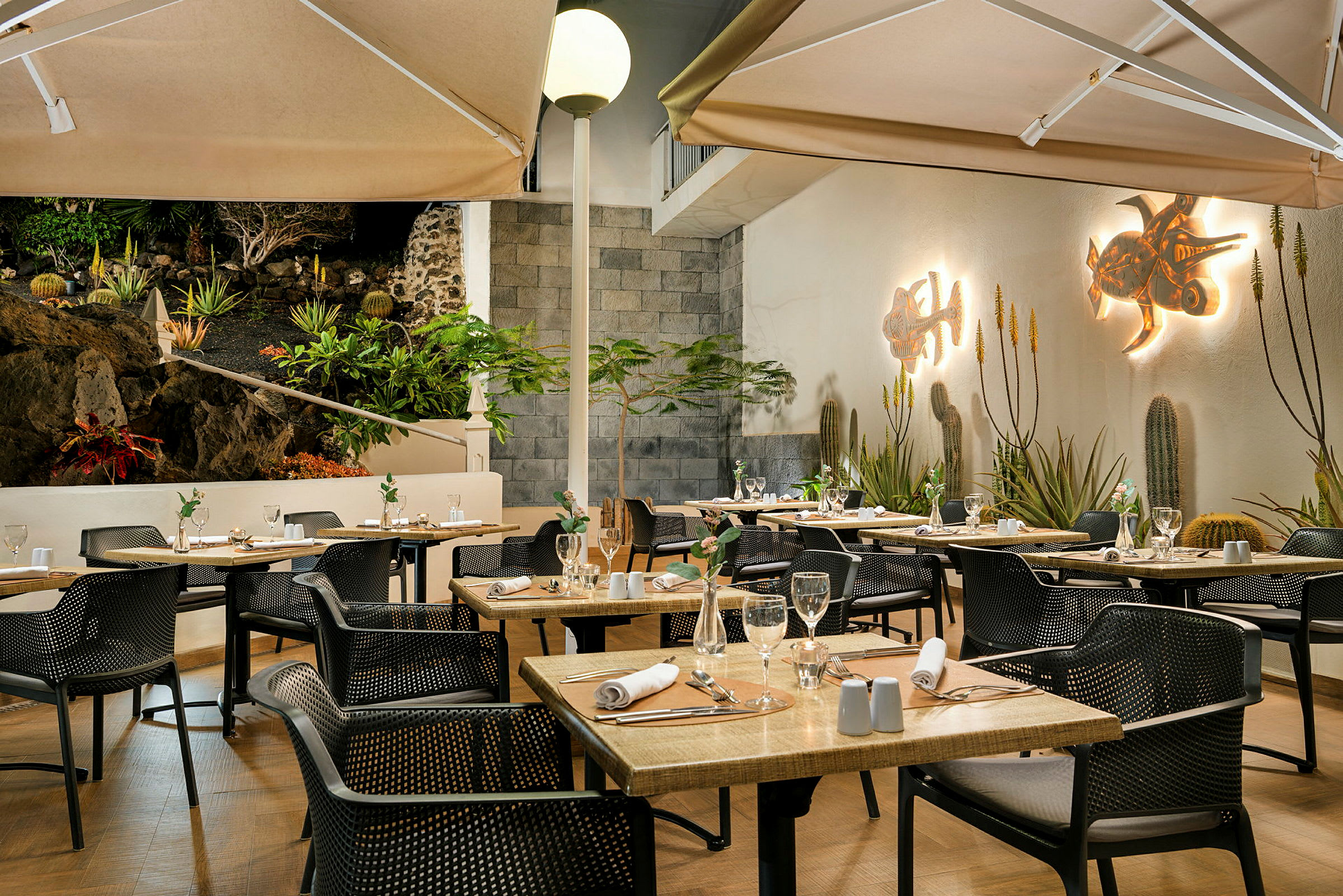 En godt opplyst restaurantterrasse med dekkede bord og stoler. Veggene er dekorert med kunst og omgivelsene har planter og en steinmur. (AI-generert tekst)