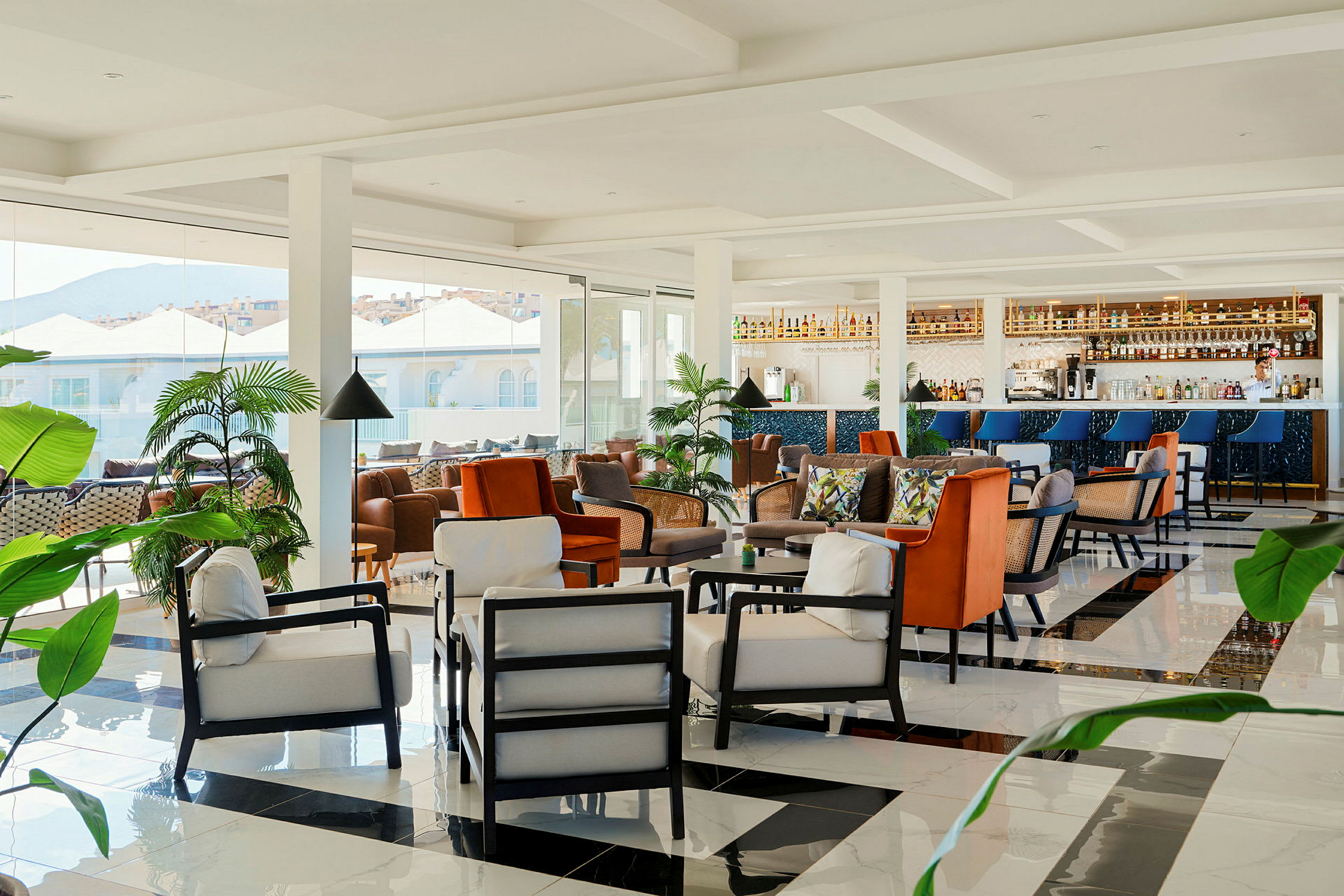 En moderne lounge med komfortable sittemøbler, en bar og frodige planter, samt store vinduer med utsikt mot bygninger og fjell. (AI-generert tekst)