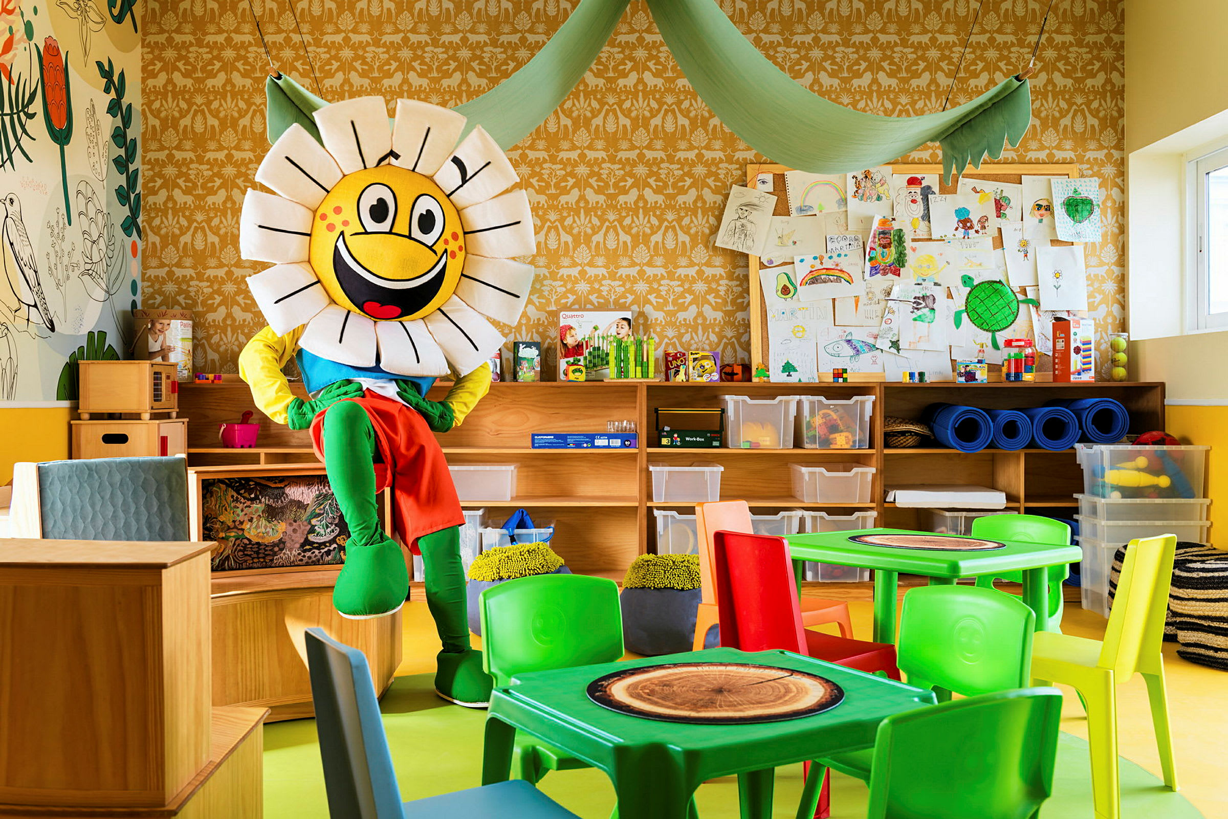 Et fargerikt rom med en maskot i form av en blomst som sitter ved møbler. Bakgrunnen viser hyller, oppbevaringsbokser og tegninger. (AI-generert tekst)
