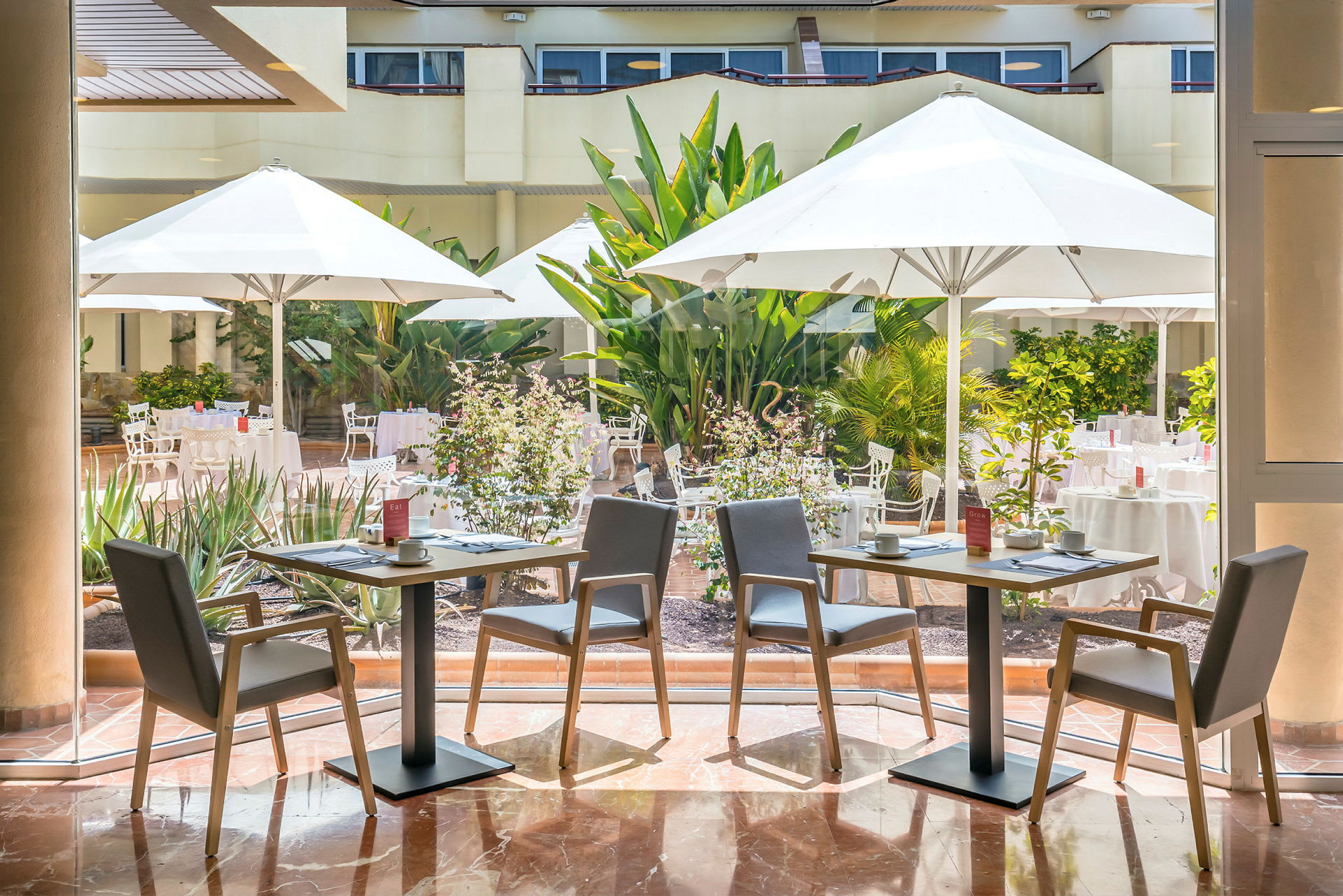 To bord med grå stoler og dekket servise står inne foran en glassvegg. Utenfor er det en terrasse med hvite parasoller, bord og stoler omgitt av frodig vegetasjon. (AI-generert tekst)