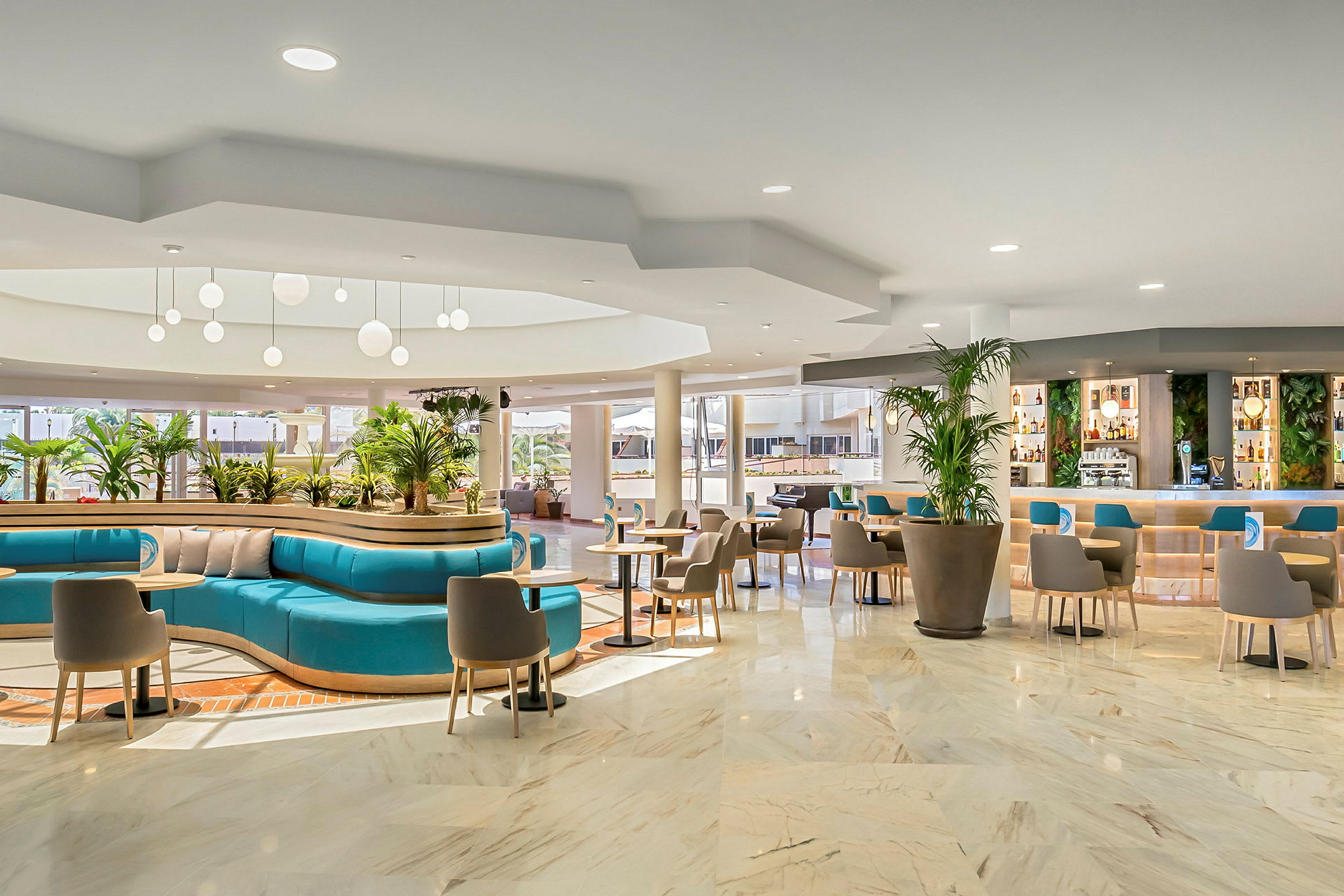 Et moderne hotellområde med sittegrupper, turkise sofaer og store potteplanter. Taklamper henger fra taket, og en innbydende bar med hyller er synlig i bakgrunnen. (AI-generert tekst)