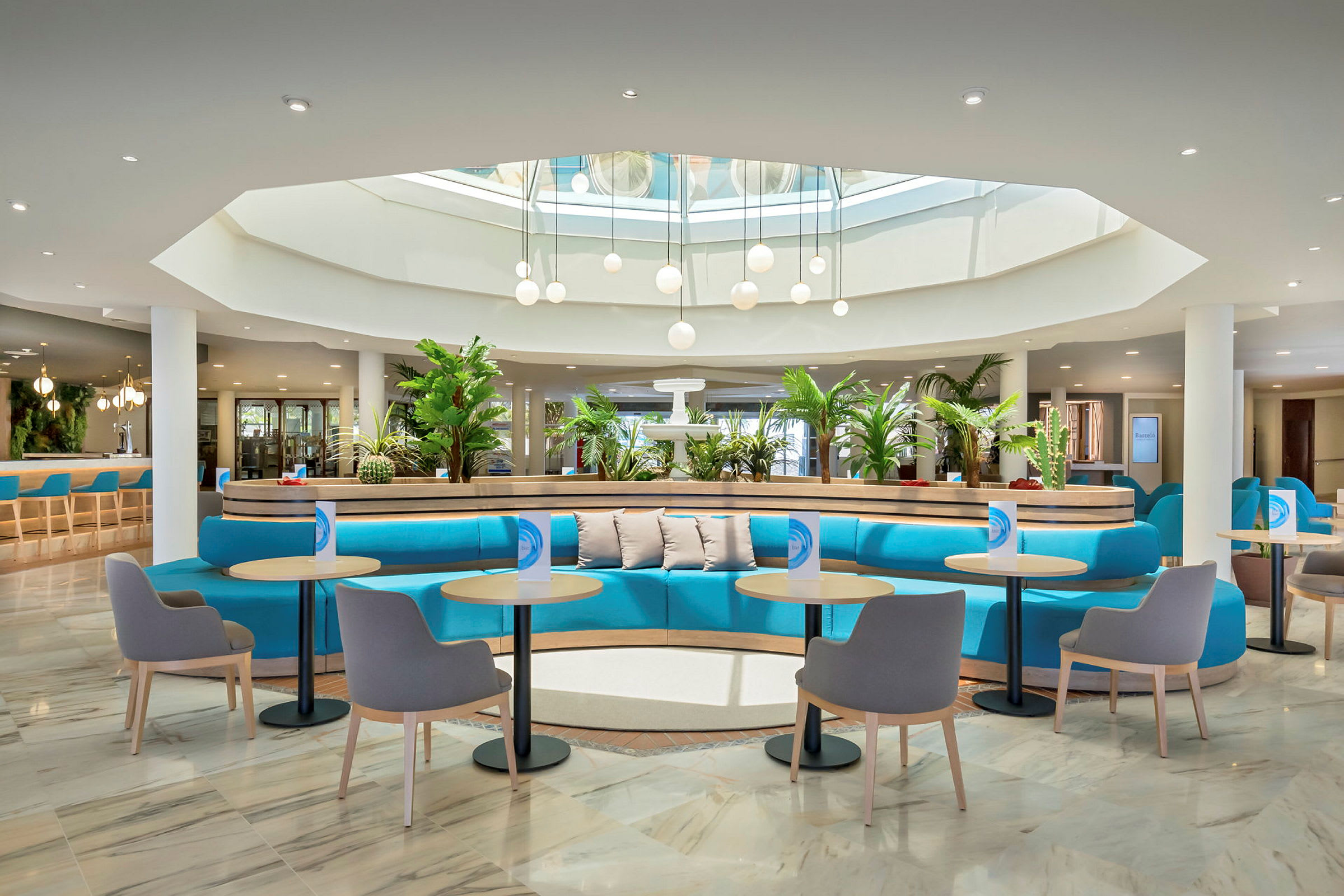 En moderne hotellobby med blå sittebenker, små bord og stoler. Planter pynter området, og takbelysning med glasskupler vises under et takvindu. (AI-generert tekst)