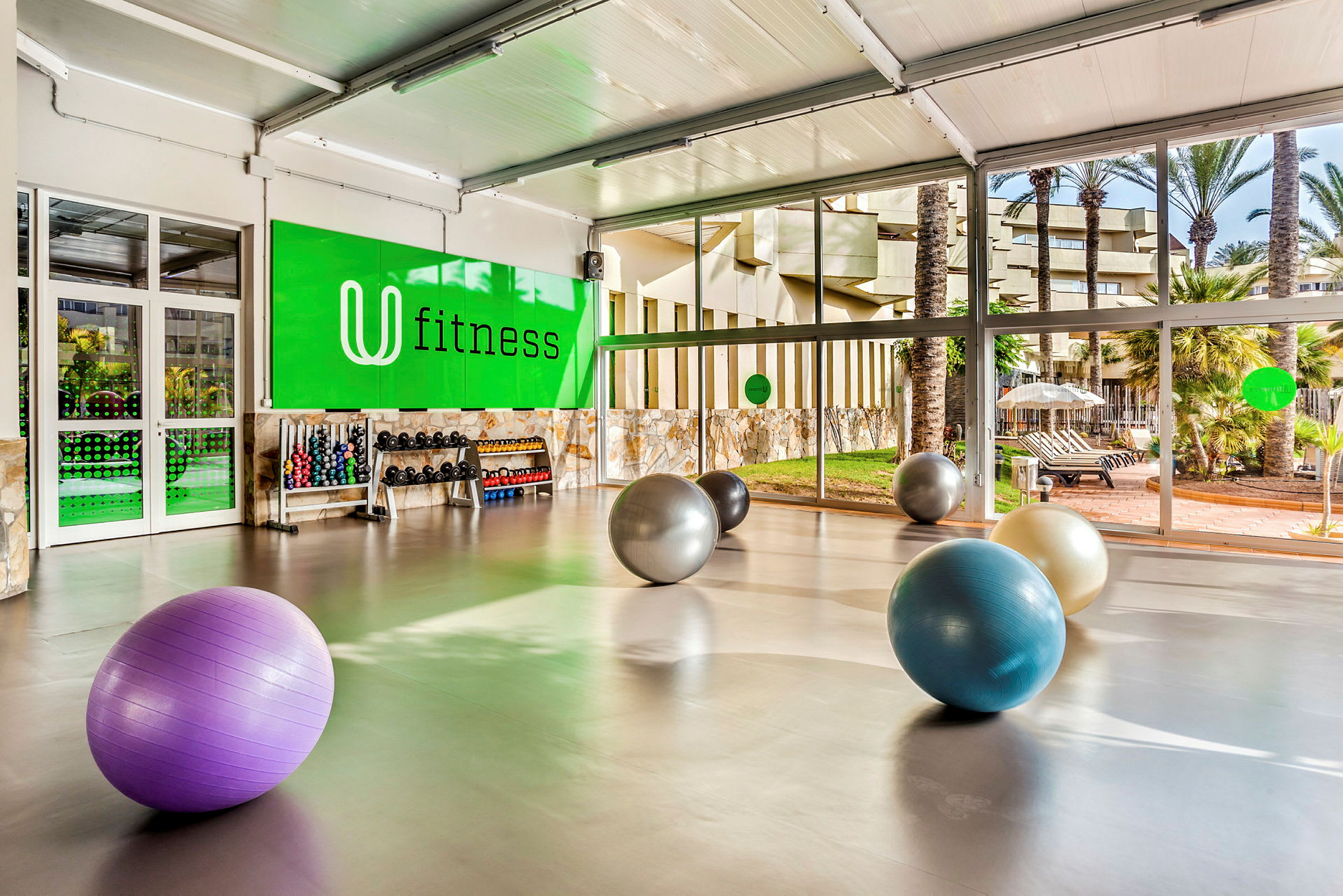 Et treningssenter med store vinduer og treningsballer på gulvet. Håndvektsstativ og treningsutstyr ses mot en grønn vegg merket "fitness". (AI-generert tekst)