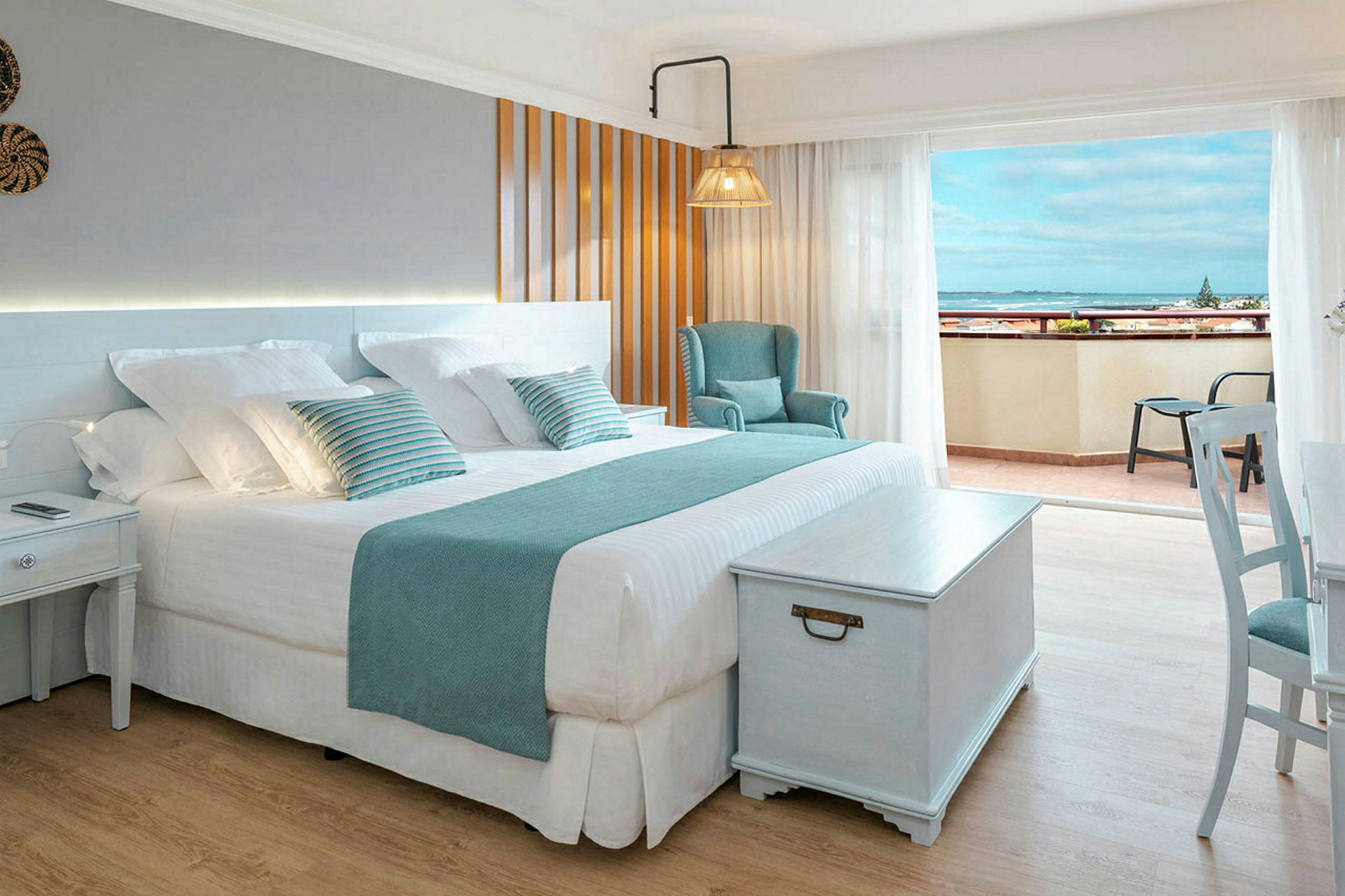 Et lyst hotellrom med en stor dobbeltseng, stol og et bord, samt en balkong med utsikt over havet. (AI-generert tekst)