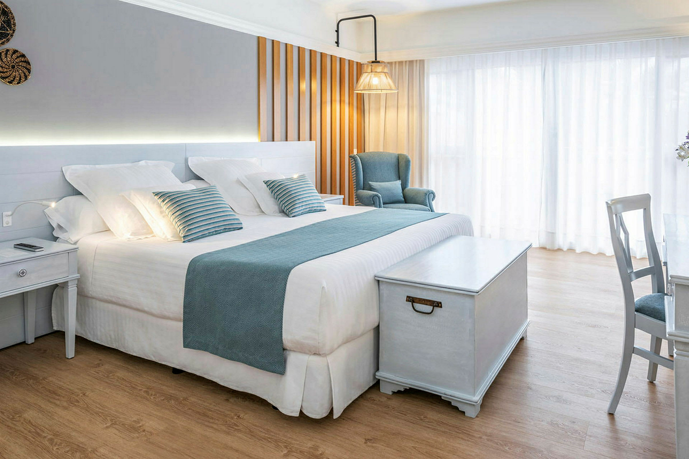 Et lyst og elegant hotellrom med en stor seng, blå dekorasjoner, en lenestol og et trebord. (AI-generert tekst)