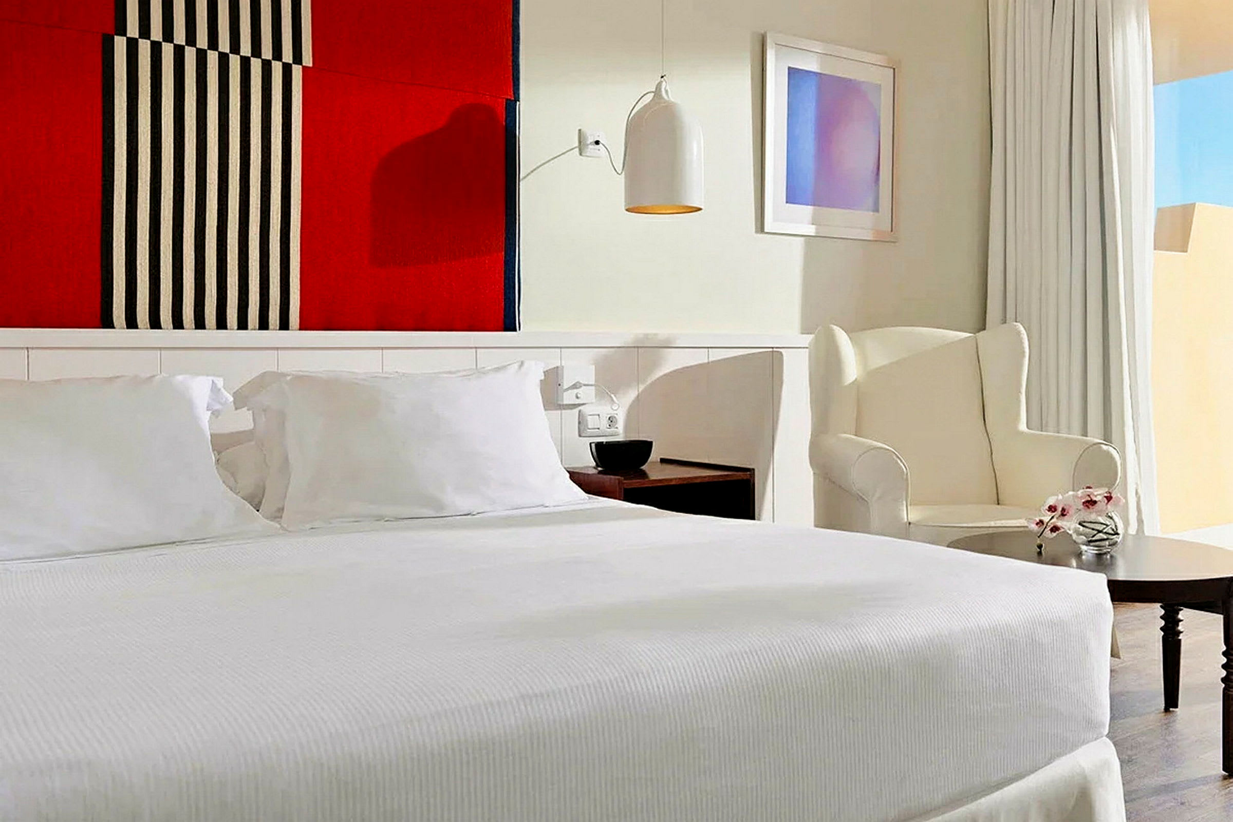 Et stilrent hotellrom med en stor seng og hvitt sengetøy, en lenestol ved siden av et lite bord, samt en fargerik tekstil veggdekorasjon over sengen. (AI-generert tekst)