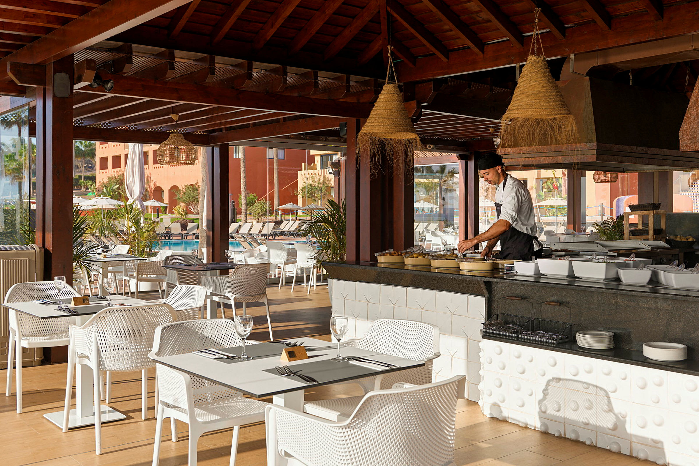 En restaurant med tremøbler på en terrasse. Bordene og stolene er hvite, og en kokk er ved matstasjonen i midten. (AI-generert tekst)