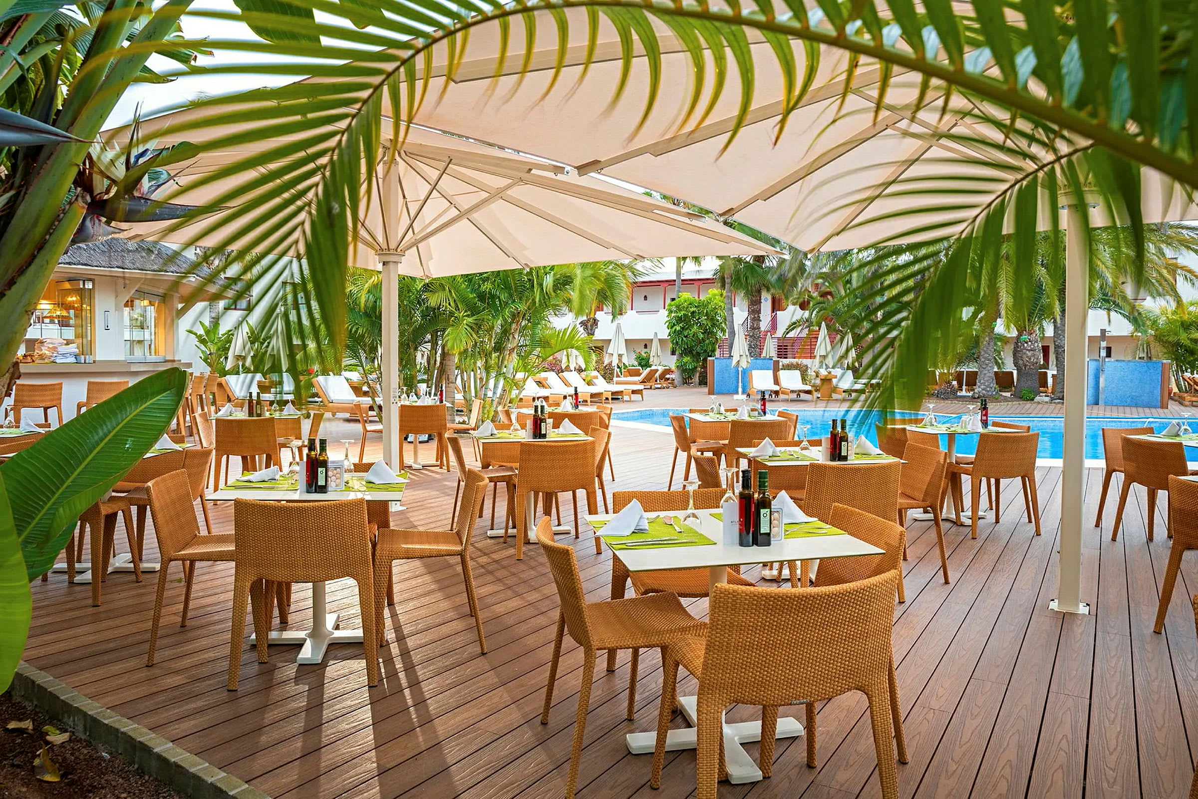 Utendørsrestaurant med tredekke, bord med duker og stoler, omgitt av grøntområder og et svømmebasseng. (AI-generert tekst)
