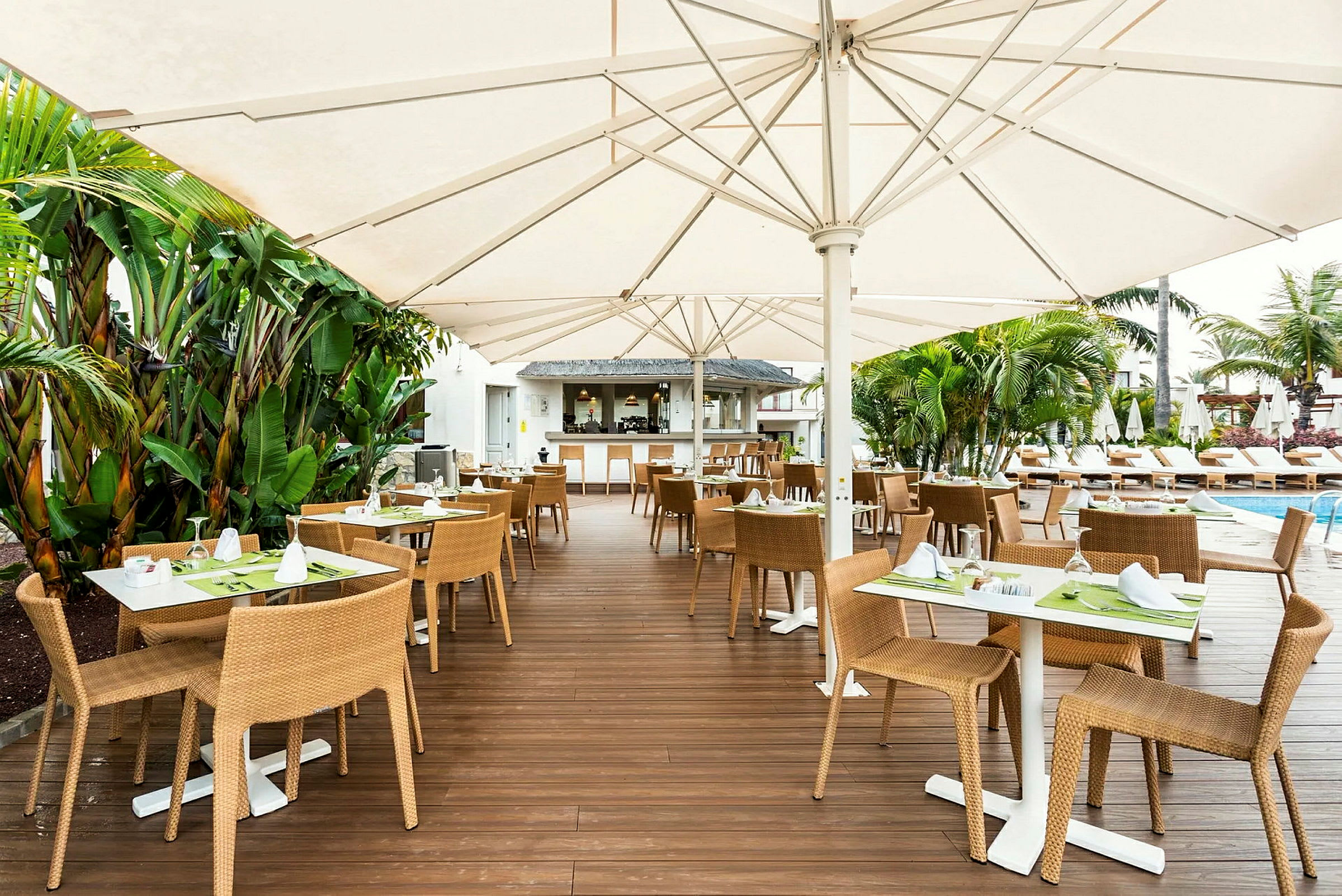Utendørsservering med bord og stoler plassert på en treterrasse, omgitt av grønne planter og med solsenger ved et basseng i bakgrunnen. (AI-generert tekst)