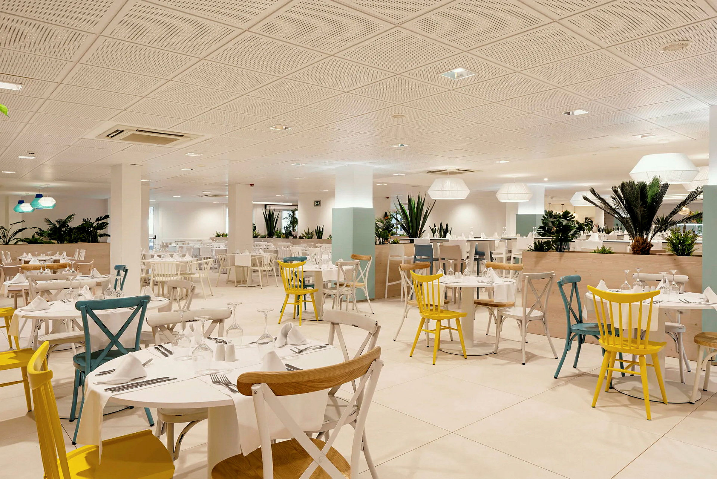Interiøret til en lys restaurant med mange bord og stoler i ulike farger, dekorert med grønne planter. (AI-generert tekst)