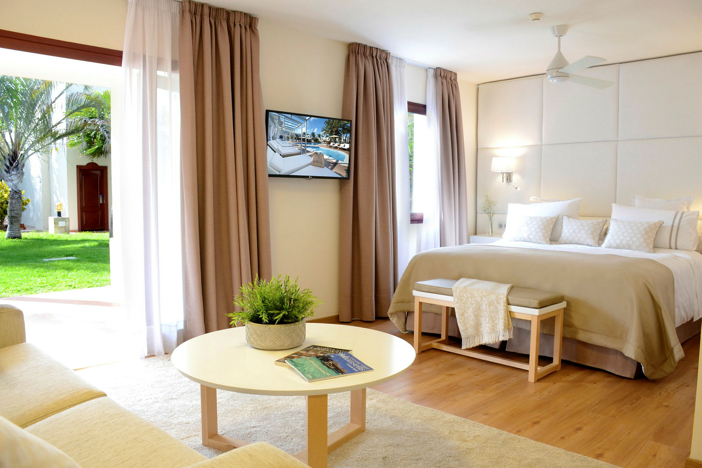 Et lyst hotellrom med en stor seng, veggmontert TV og en sittegruppe foran et rundt bord. Store gardiner med utsikt mot en hage. (AI-generert tekst)